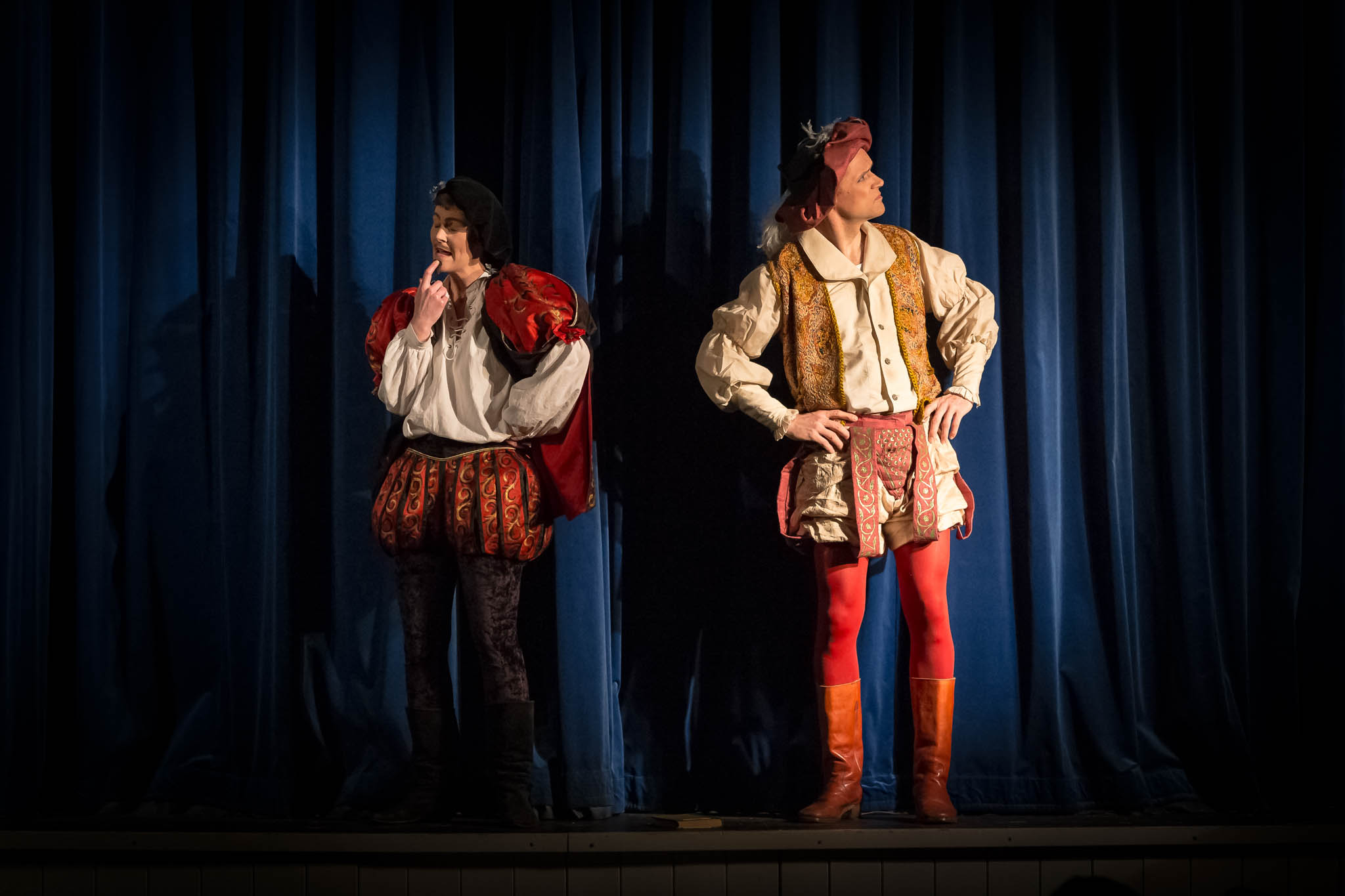 Two actors in Renaissance attire perform on stage with a blue curtain backdrop.