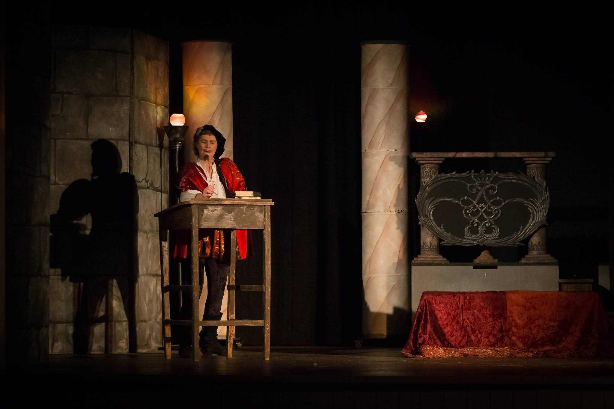Actor in period costume delivers a monologue on a dimly lit stage with stone pillars and ornate backdrop.