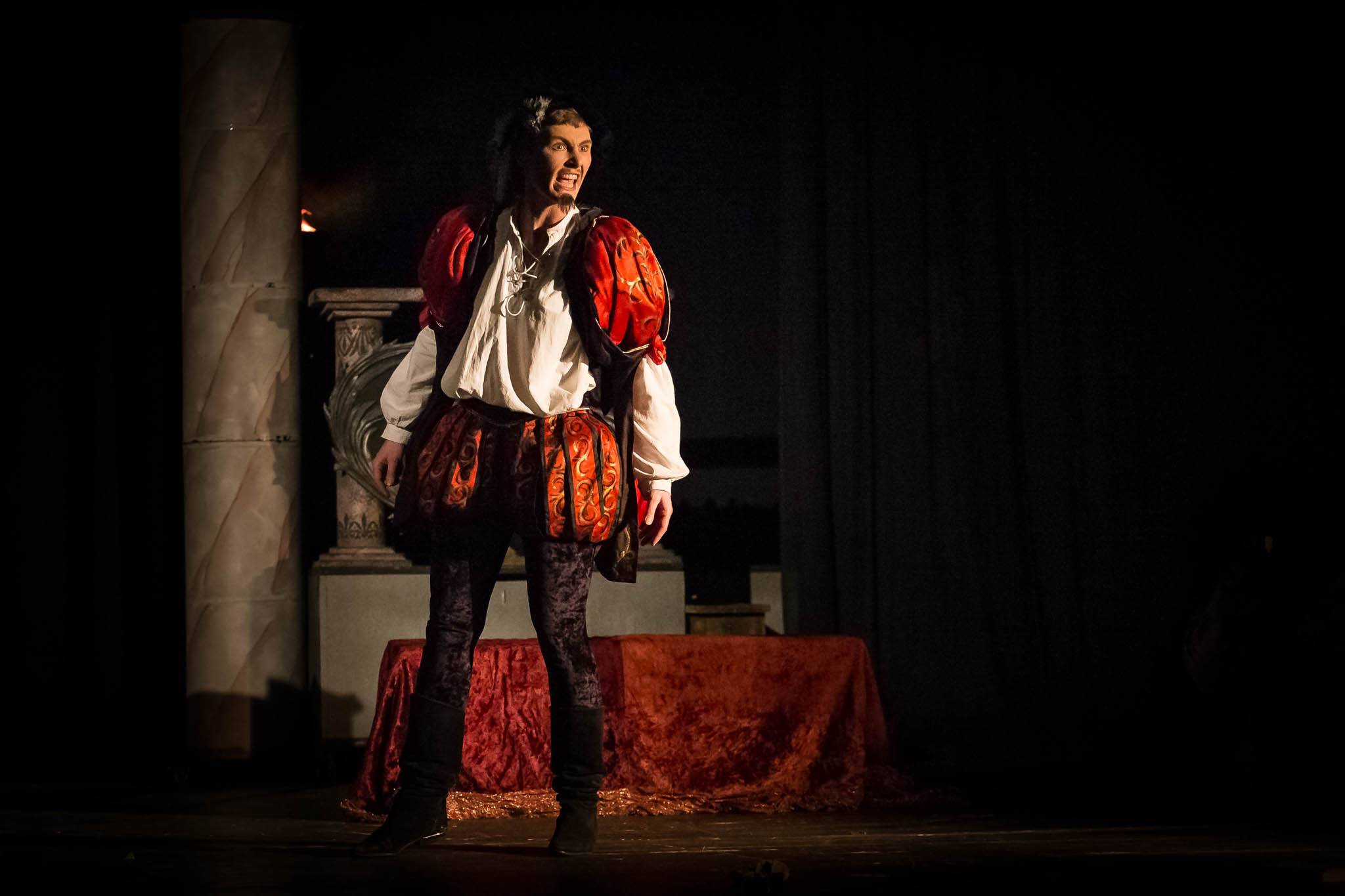 Actor in medieval costume performs passionately on stage, with dramatic lighting enhancing the scene.
