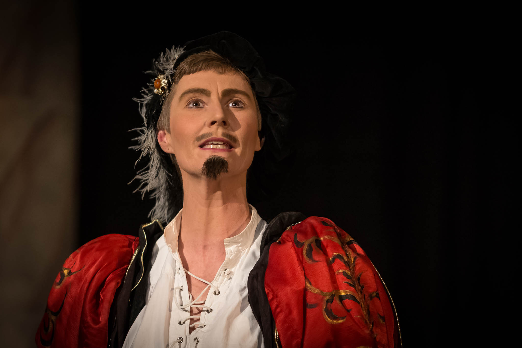 Actor in elaborate red and white Renaissance costume performing on stage with dramatic expression.