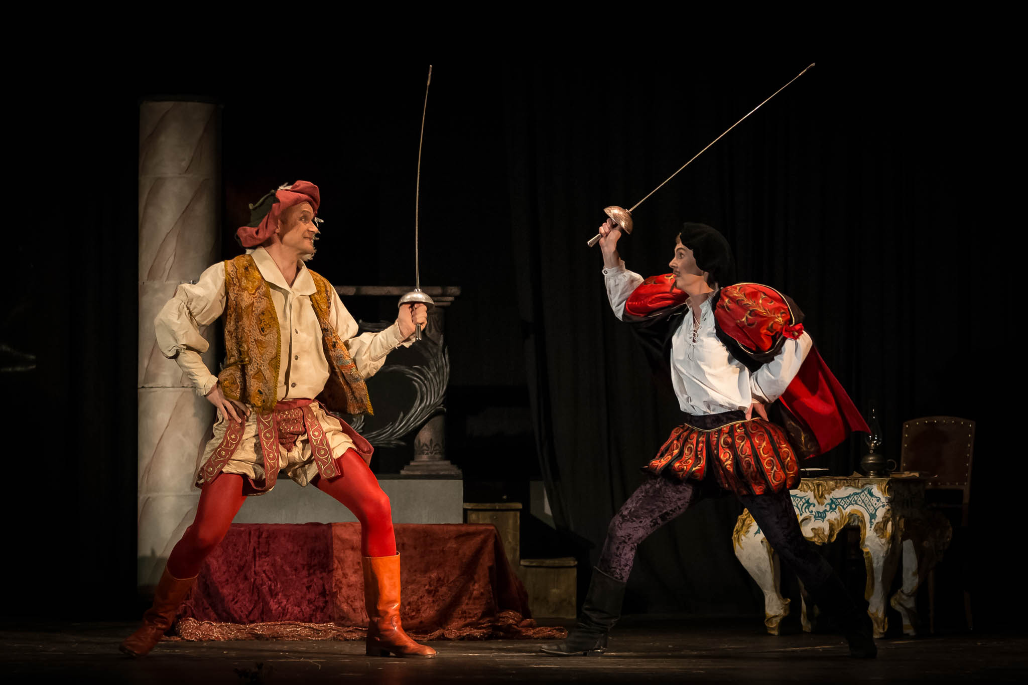 Two actors in colorful period costumes duel with swords on stage during a theatrical performance.