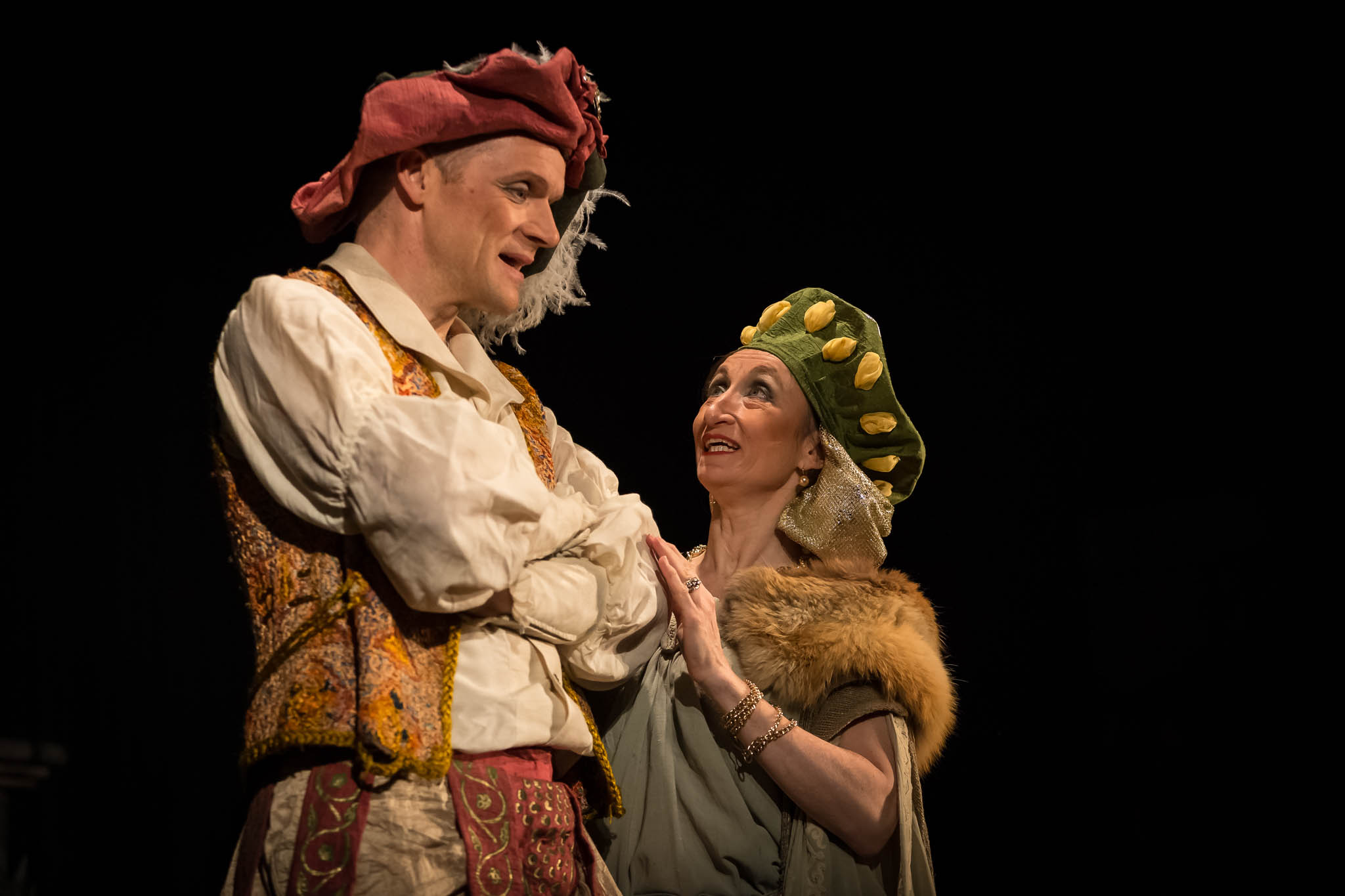 Two actors in elaborate Renaissance costumes perform on stage, a woman gazes up at a smiling man.