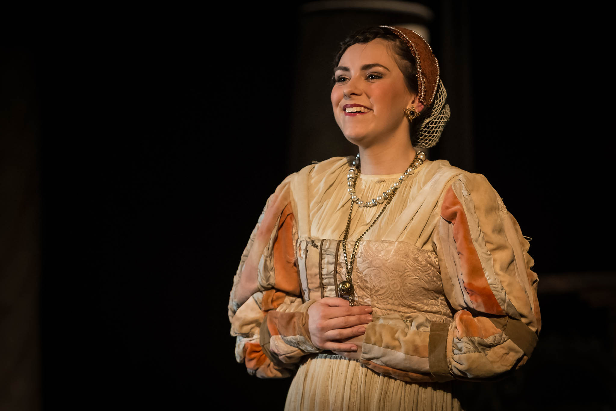 Woman in historical costume with pearl necklace smiling against a dark background.
