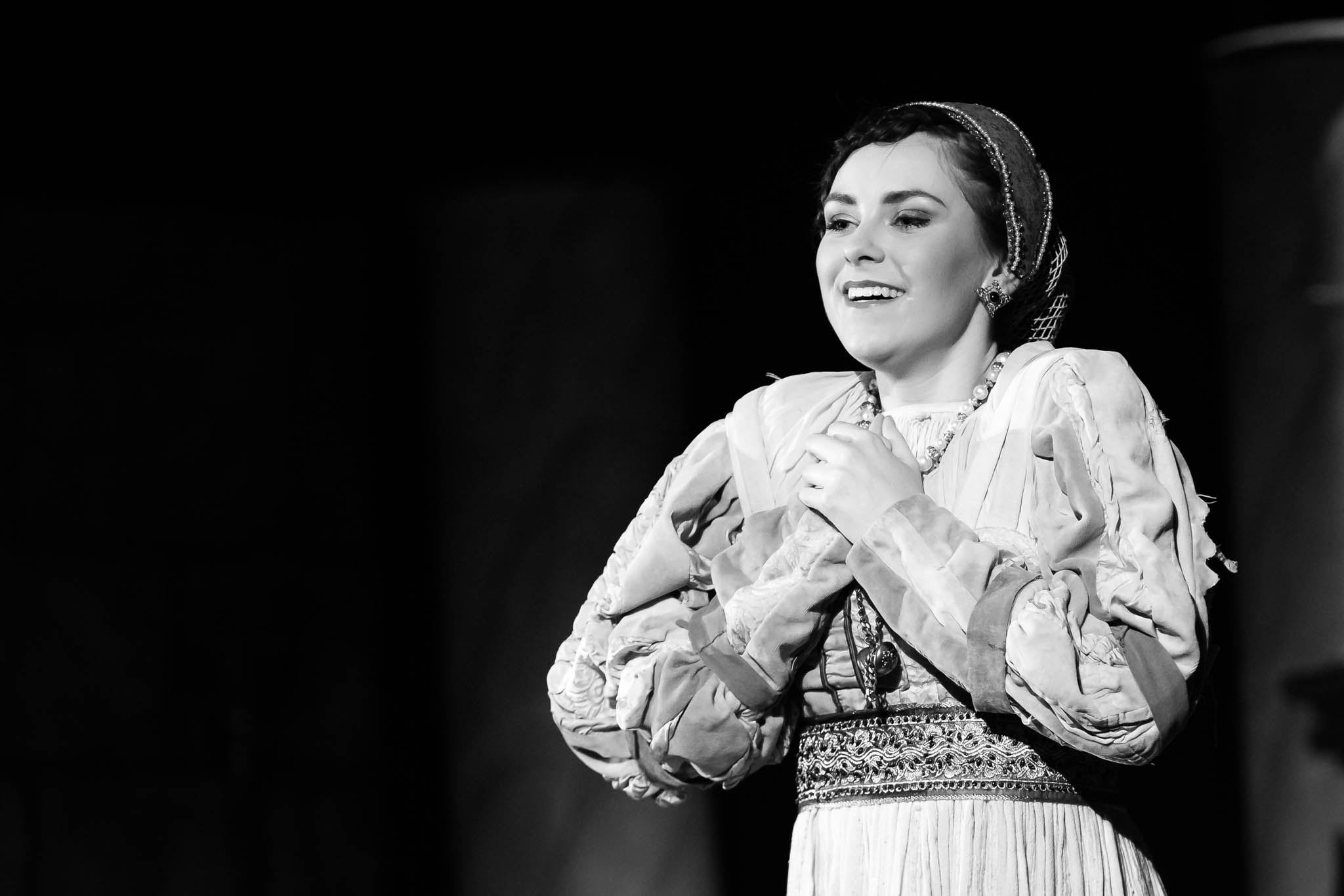 Black and white photo of a smiling woman in historical costume on stage, hands clasped, expressing joy.