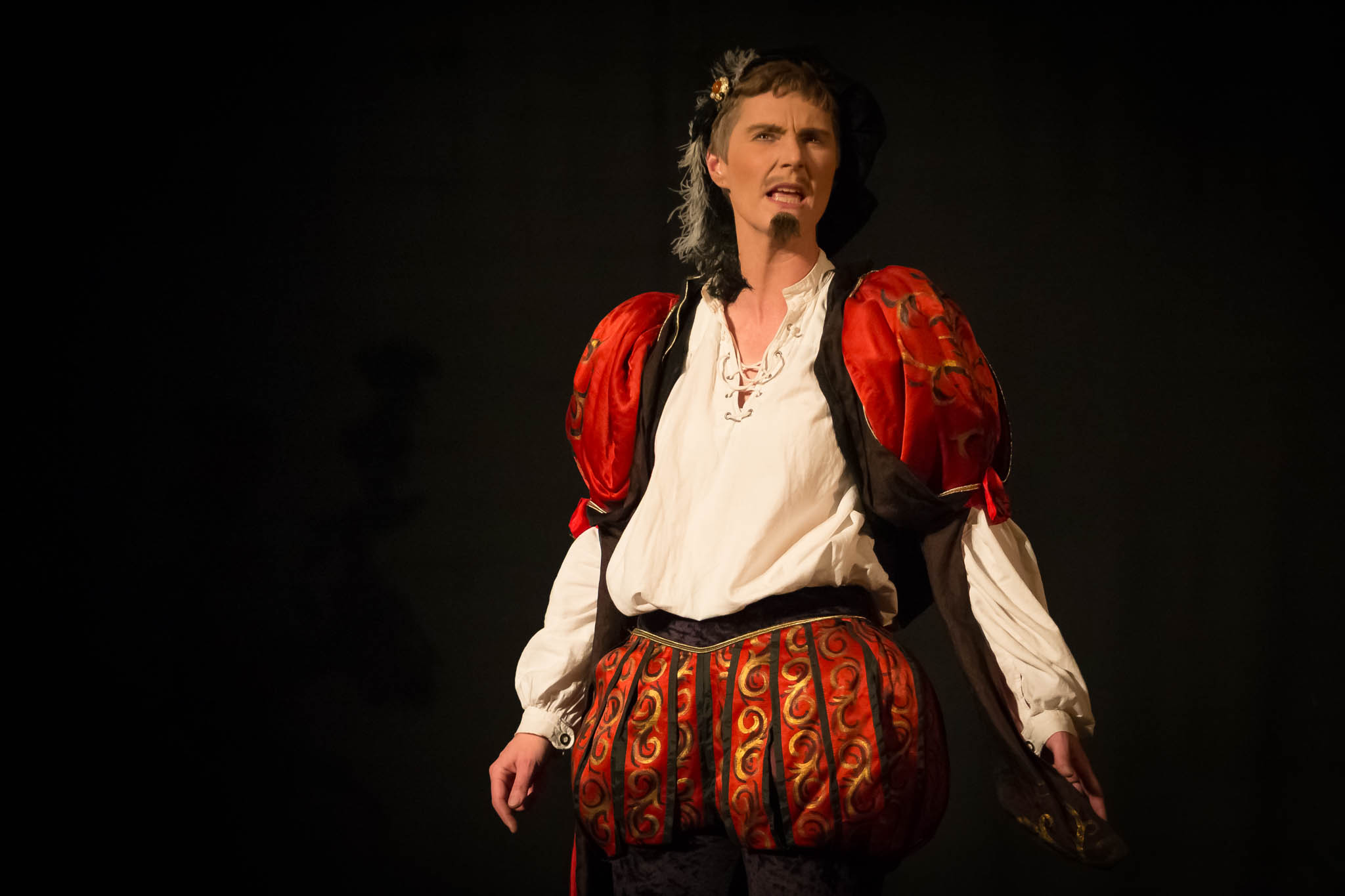 Actor in vibrant Renaissance costume performs passionately on stage against a dark background.