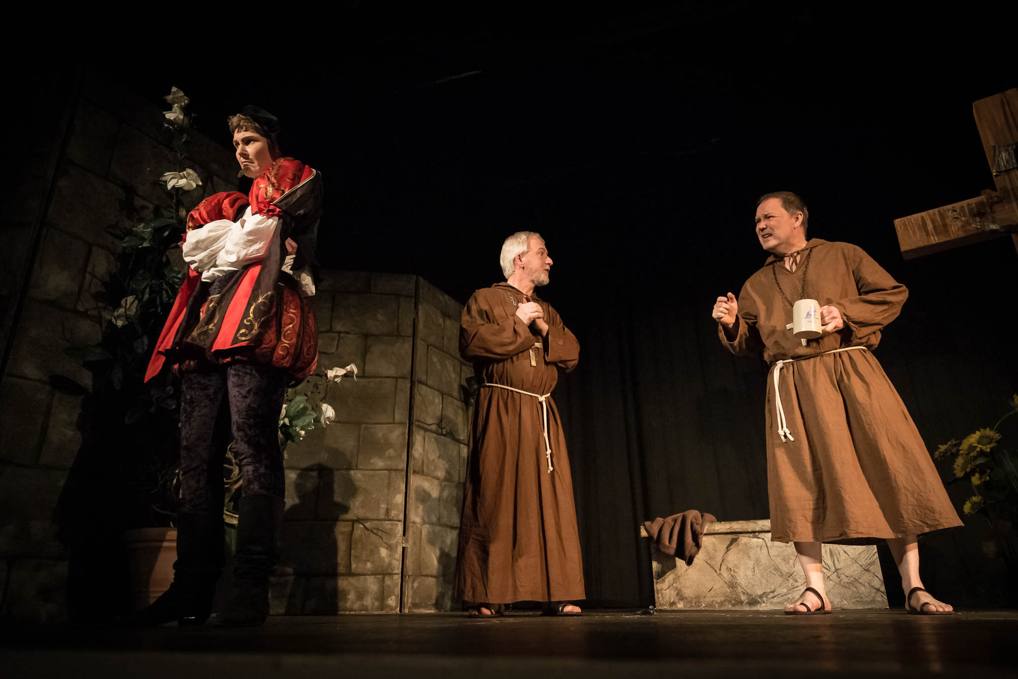 Actors perform a medieval play, featuring a nobleman and two monks in brown robes on stage with stone wall backdrop.
