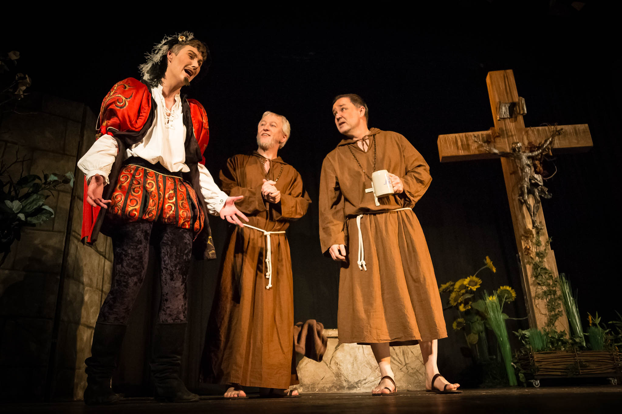 Three actors in medieval costumes perform on stage, one in vibrant attire, two as monks by a large cross with flowers.
