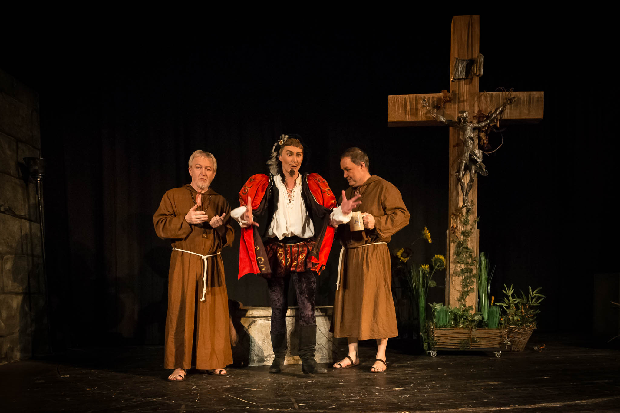 Three actors in medieval costumes perform on stage beside a large wooden cross in a theater play scene.