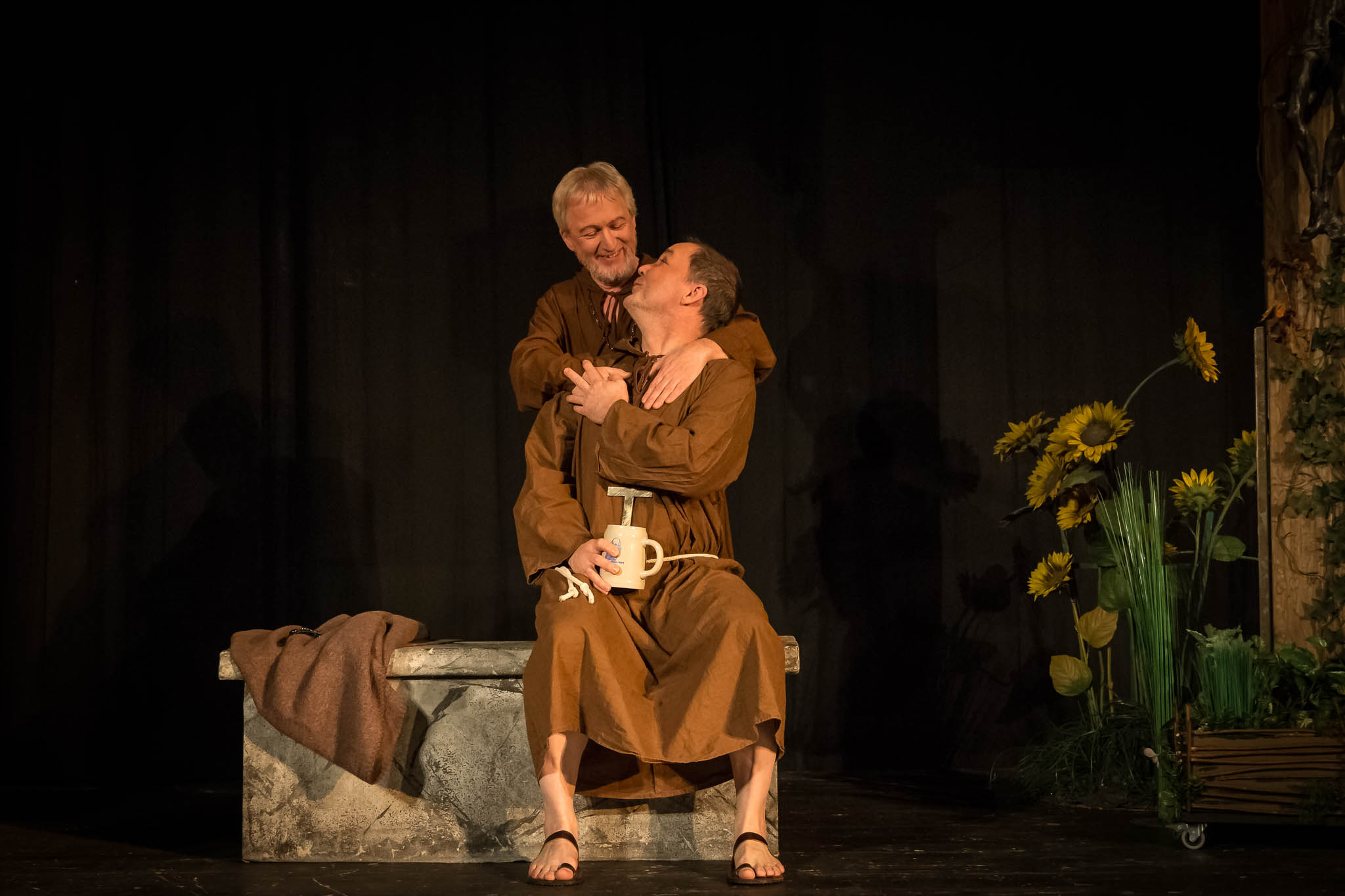 Two men in brown robes embrace on a stone bench, holding a mug, with sunflowers in the background on a dark stage.