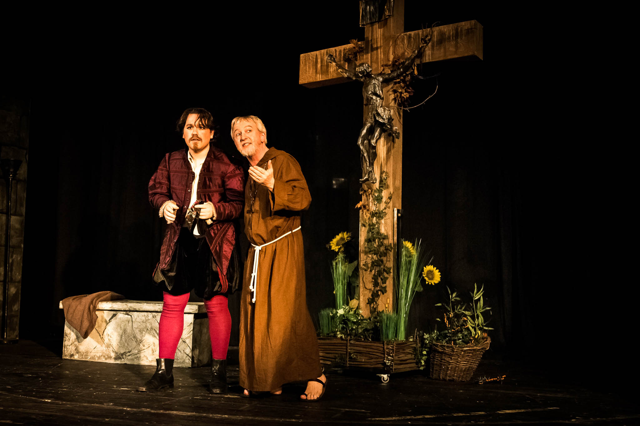 Two actors in medieval costumes perform on stage with a large cross and sunflowers in the background.