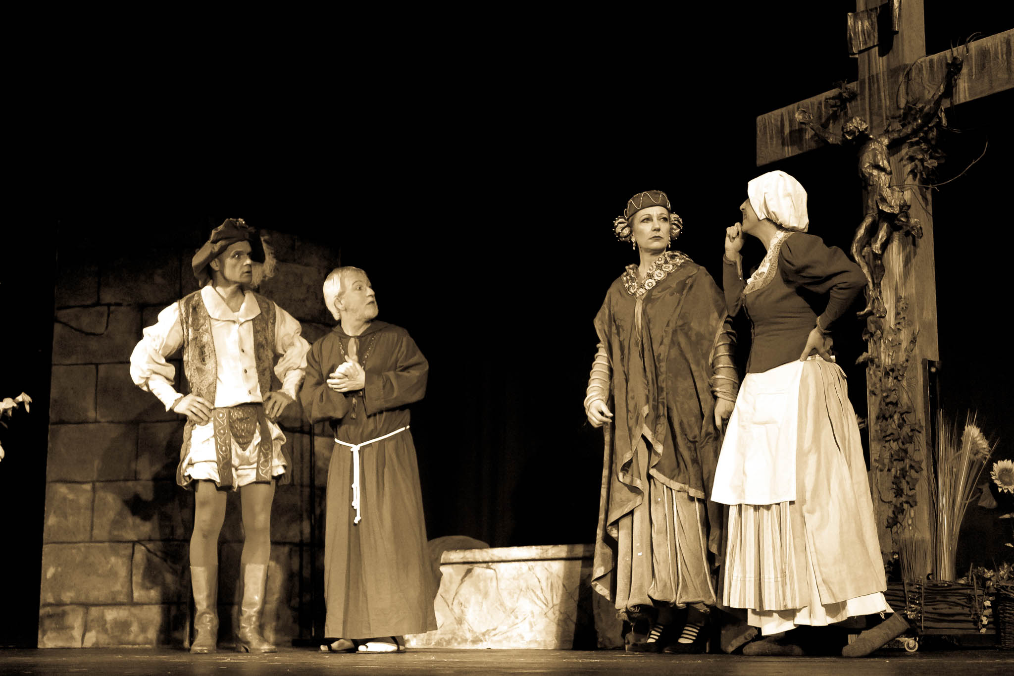 Four actors in medieval costumes perform a dramatic scene on stage, with stone walls and a large cross in the background.