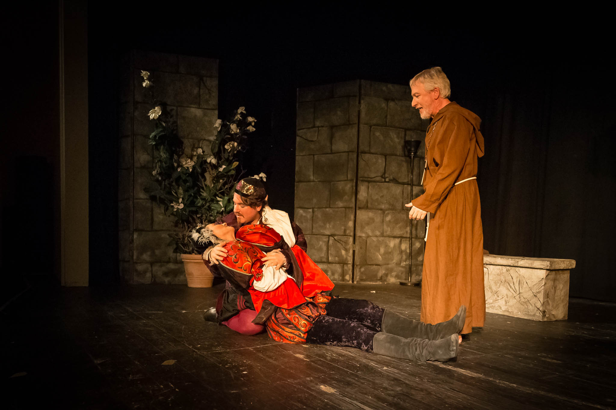 A dramatic stage scene with actors in medieval costumes, one kneeling and holding another, while a monk observes.