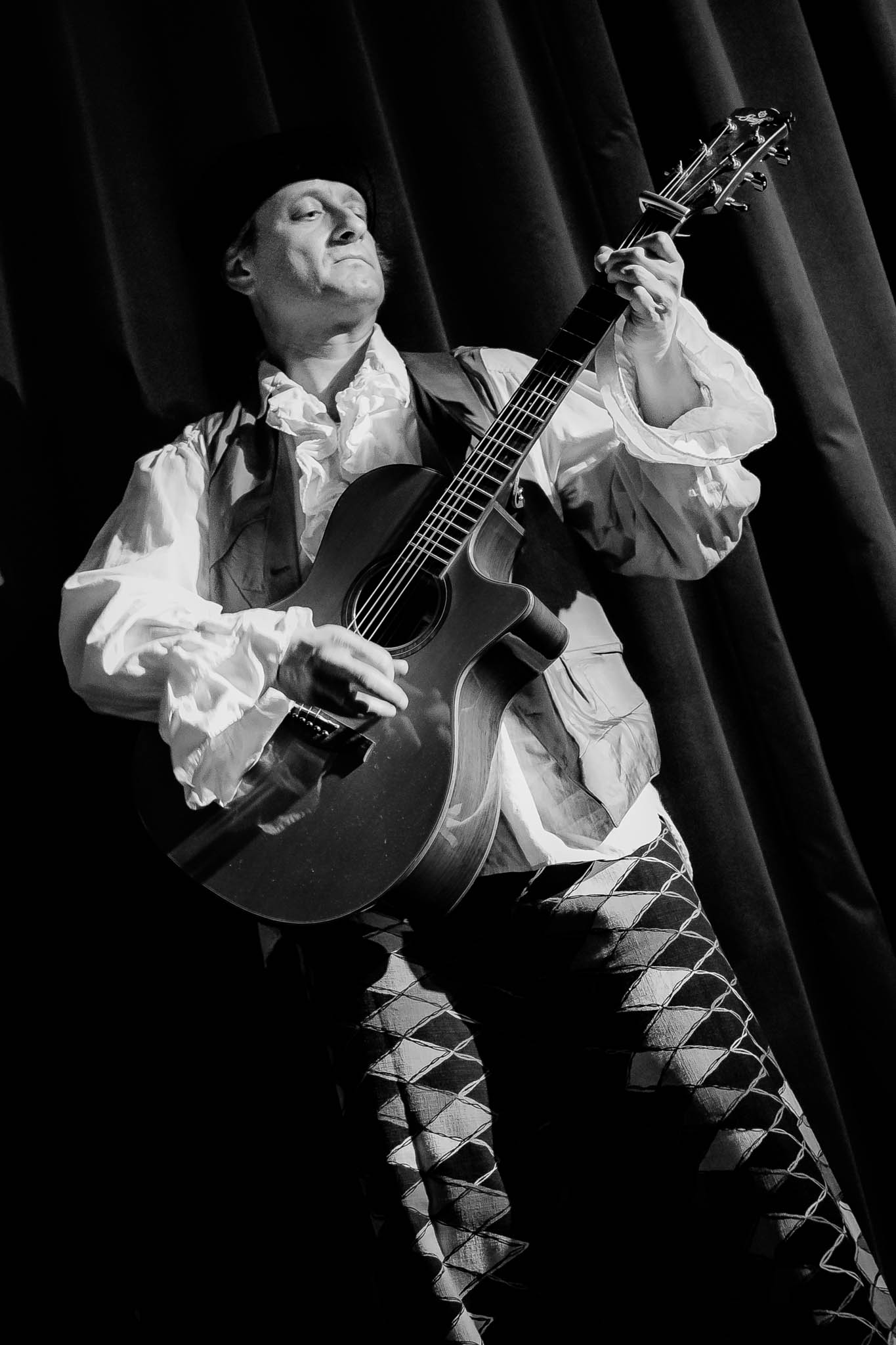 Musician in vintage costume plays acoustic guitar on stage with dramatic lighting.
