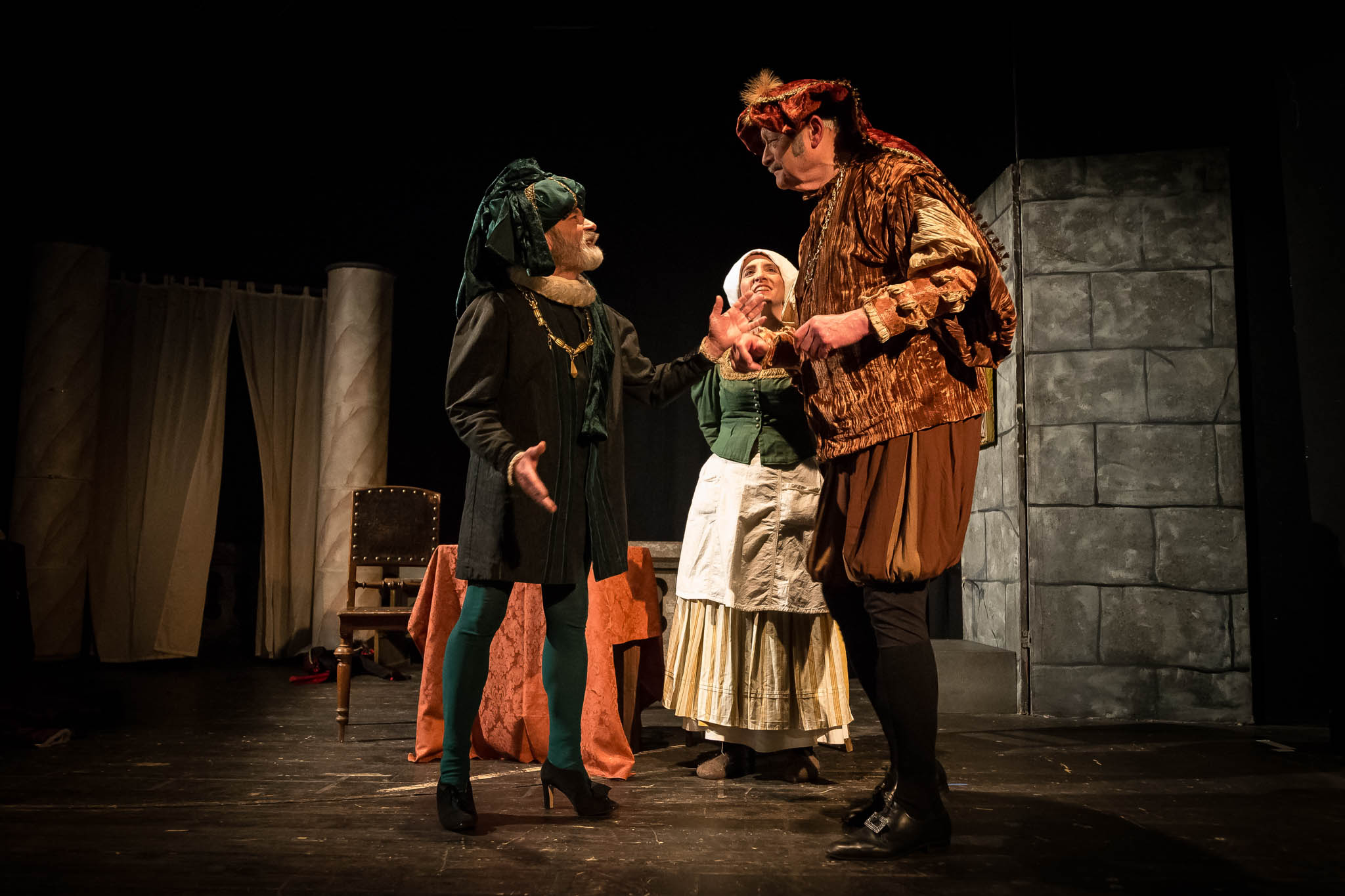 Actors in medieval costumes perform a theatrical scene on stage with dramatic lighting.