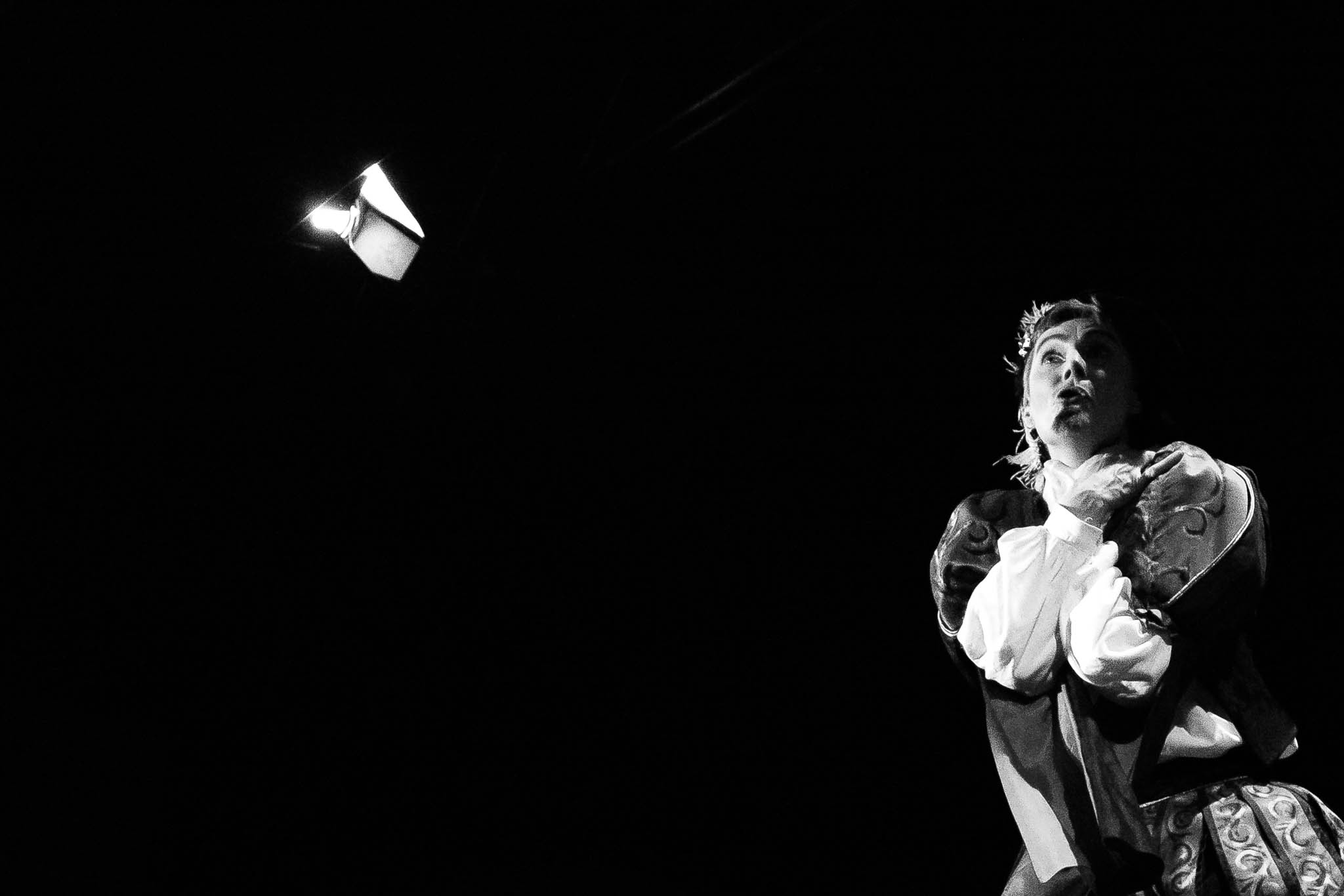 Performer in period costume dramatically poses under spotlight against dark background.