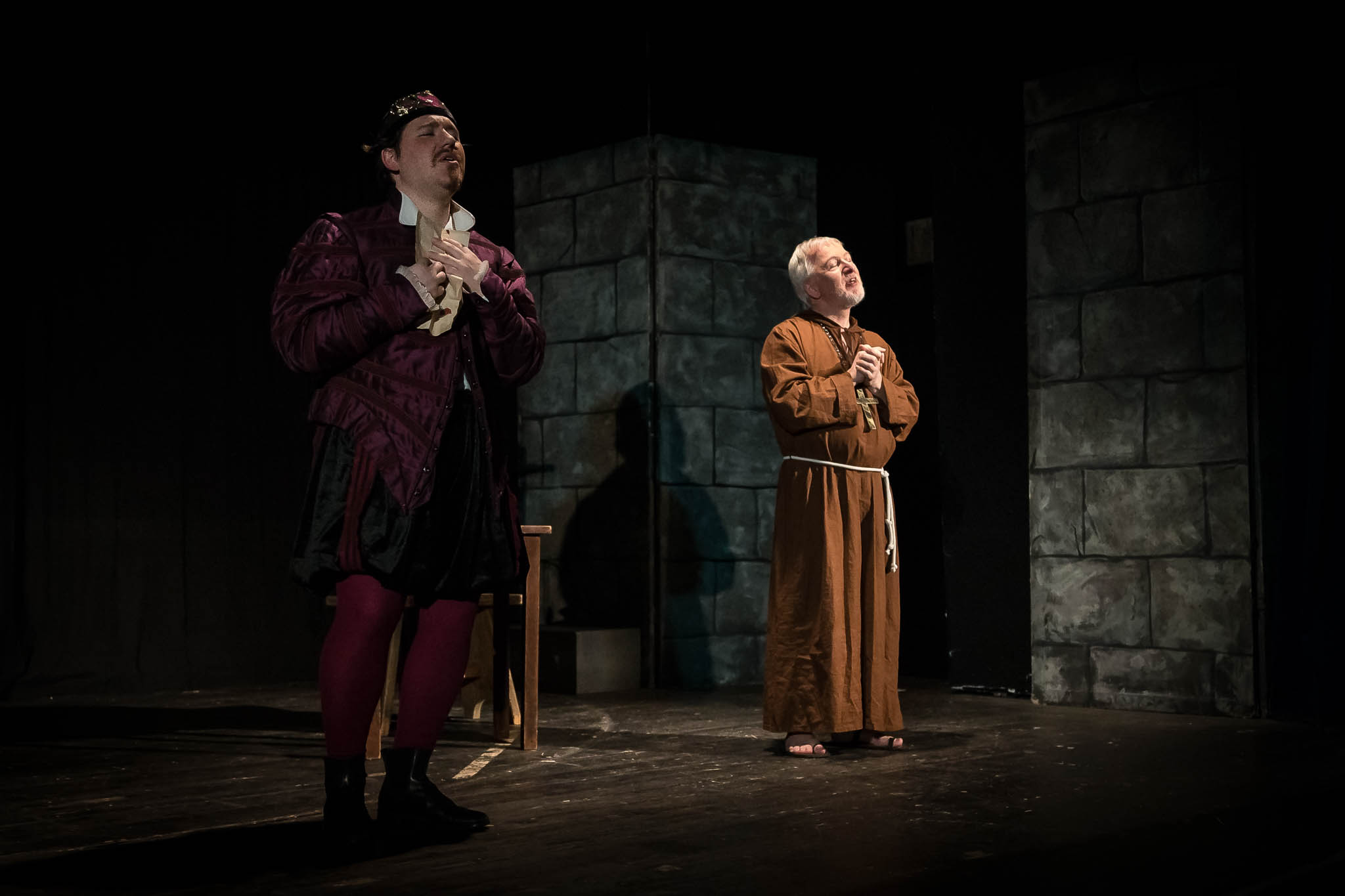 Two actors perform on stage; one in royal attire, the other dressed as a monk, against a stone wall backdrop.
