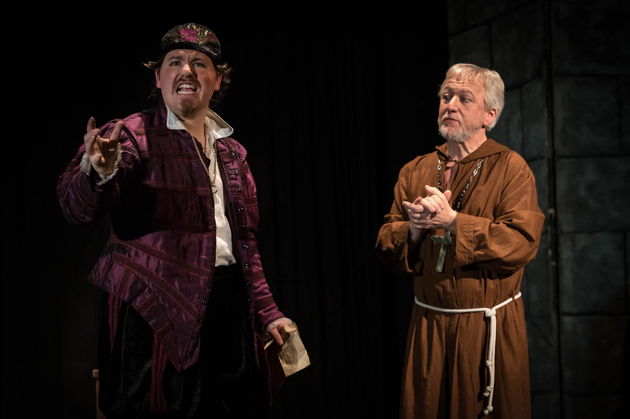 Two actors in period costumes perform on stage; one in a purple outfit gestures passionately, the other in a brown robe listens.
