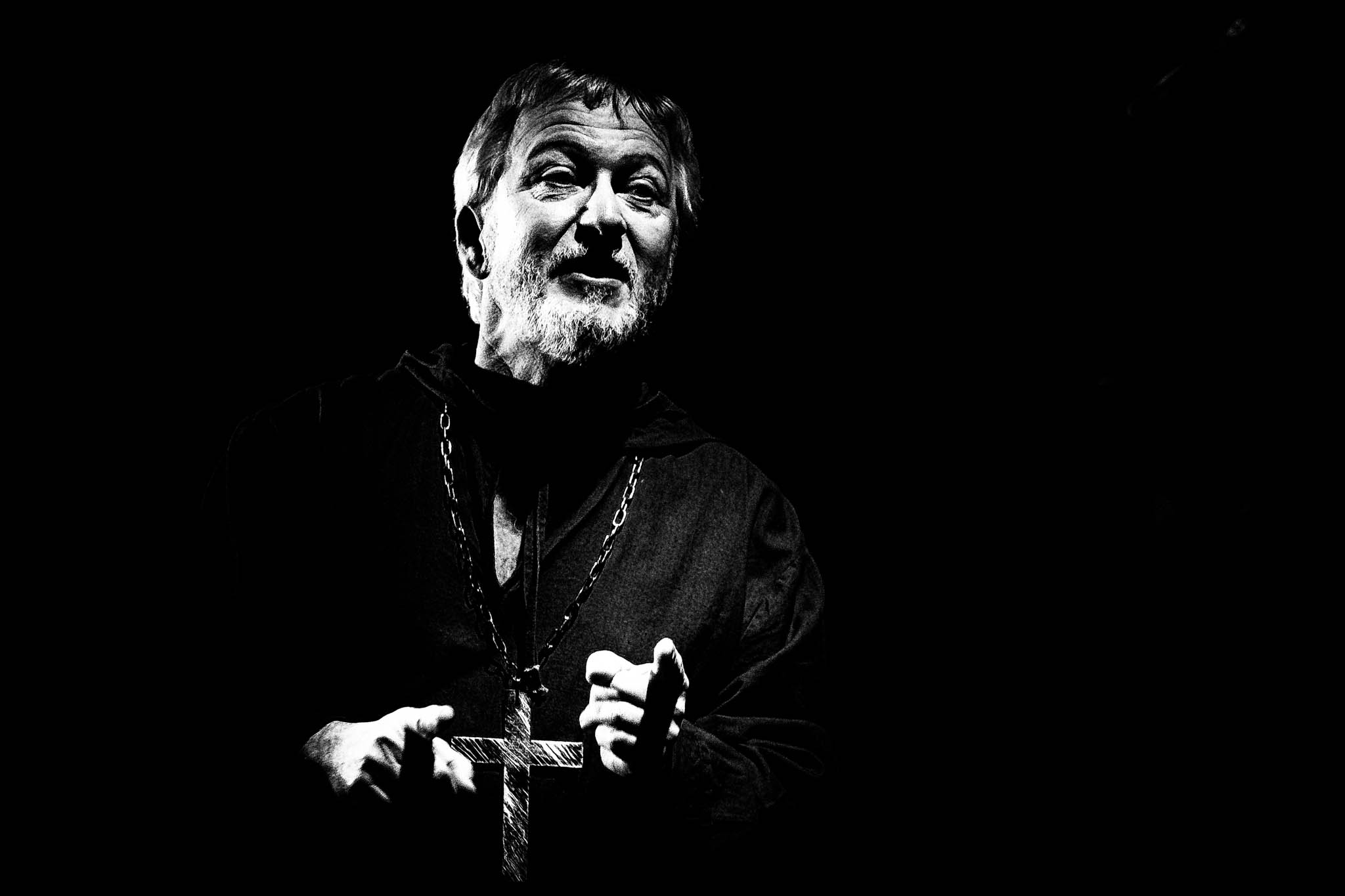 Monochrome image of a bearded man in a robe holding a large cross, set against a dark background.