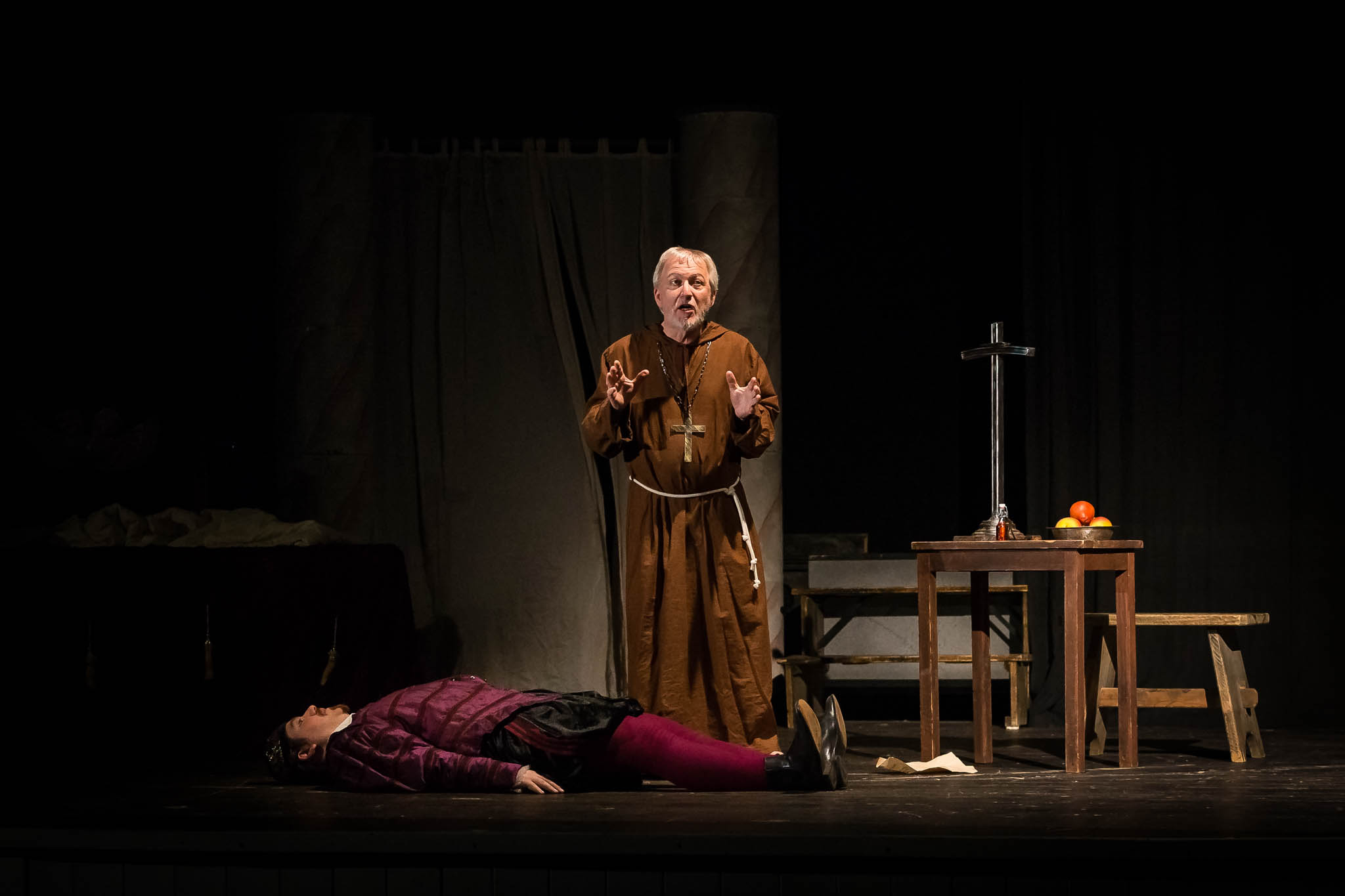 Actor in monk costume gestures beside a person lying on stage during a dramatic play scene with a rustic table.