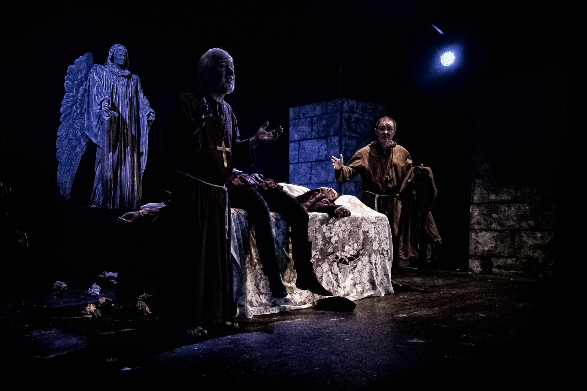 Actors perform a dramatic scene with a cross, a statue, and a figure on a bed in dim, theatrical lighting.