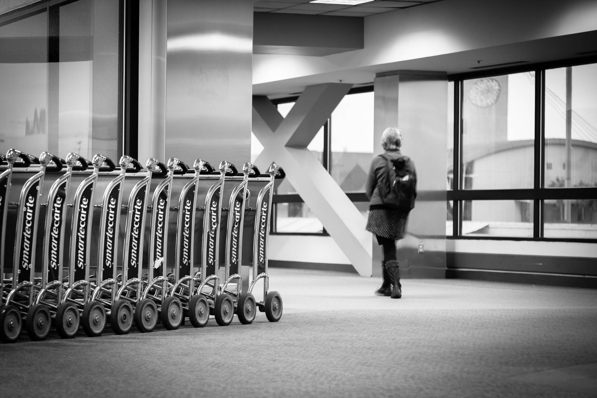 Row of airport luggage carts with a traveler walking away in a spacious, modern terminal. Black and white photo.
