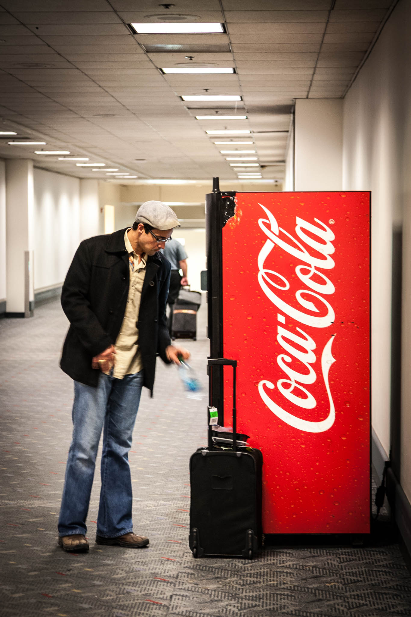 Man standing by Coca-Cola vending machine in airport corridor with luggage.