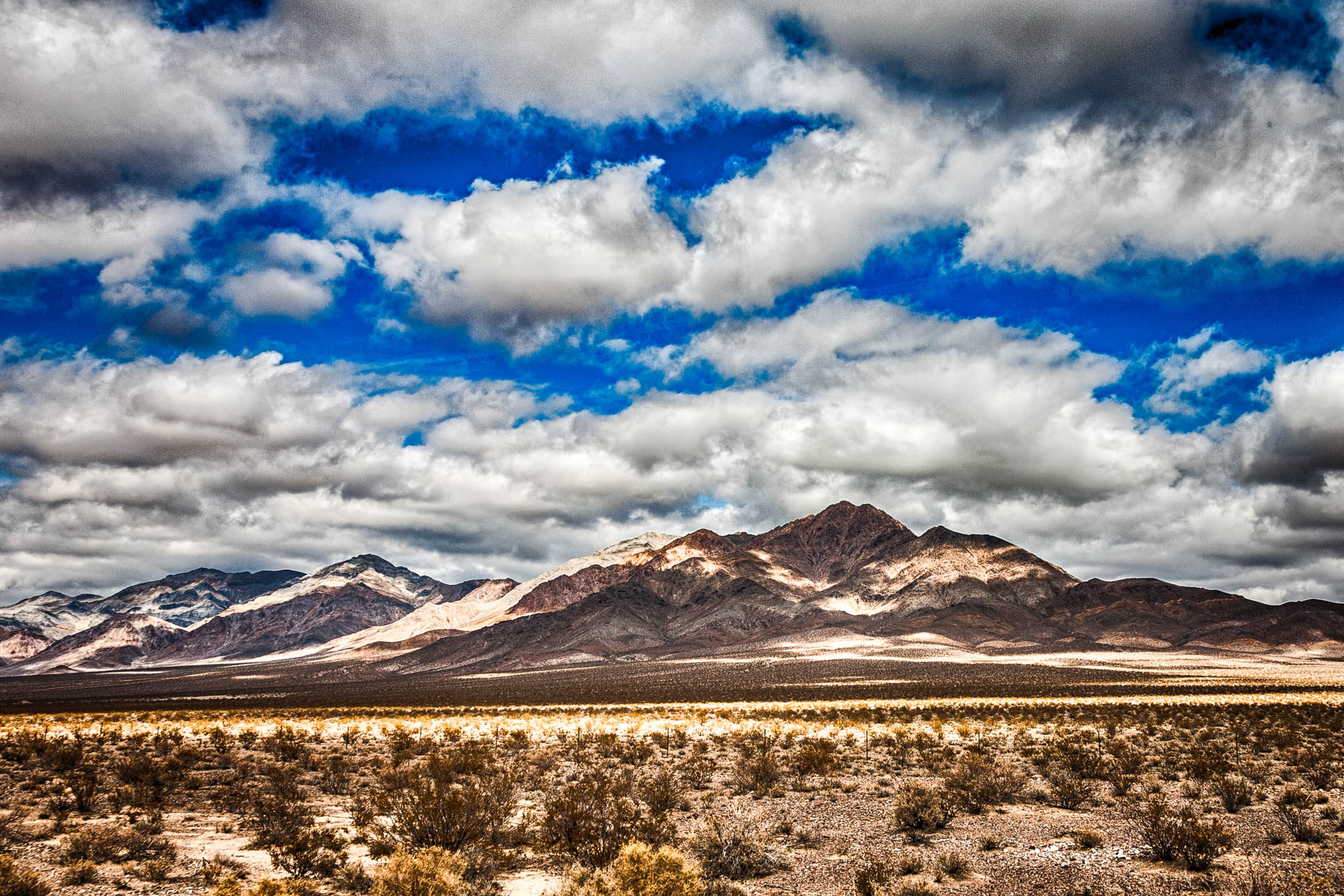 Desert landscape with rugged mountains and cloudy sky under a vibrant blue, reflecting sunlight in arid terrain.