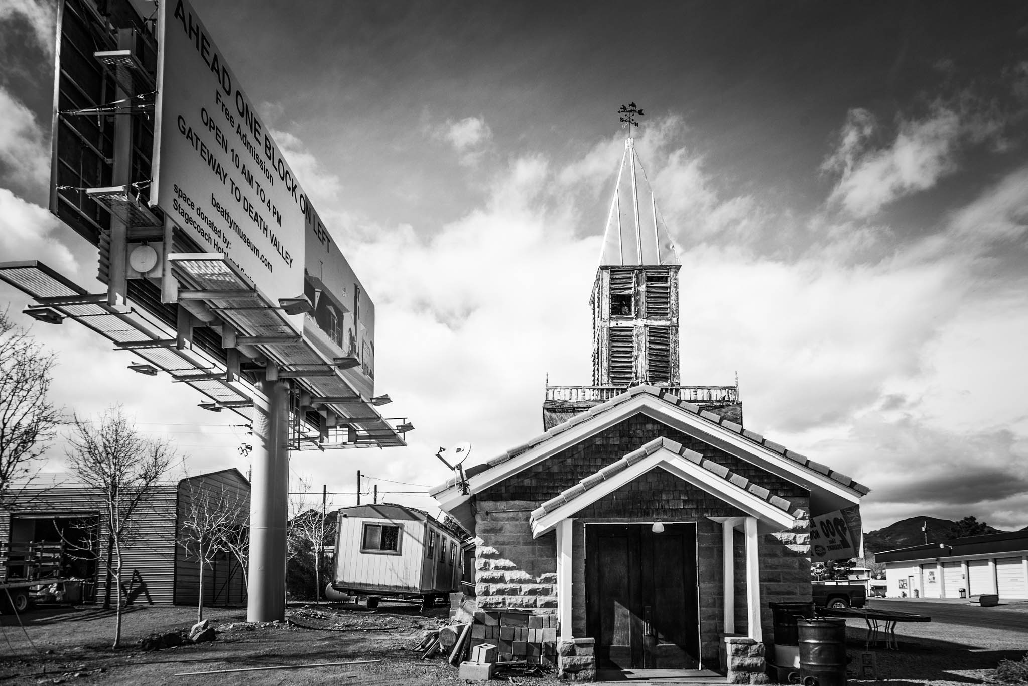 Black and white photo of a rustic church with a steeple next to a large roadside billboard under a cloudy sky.