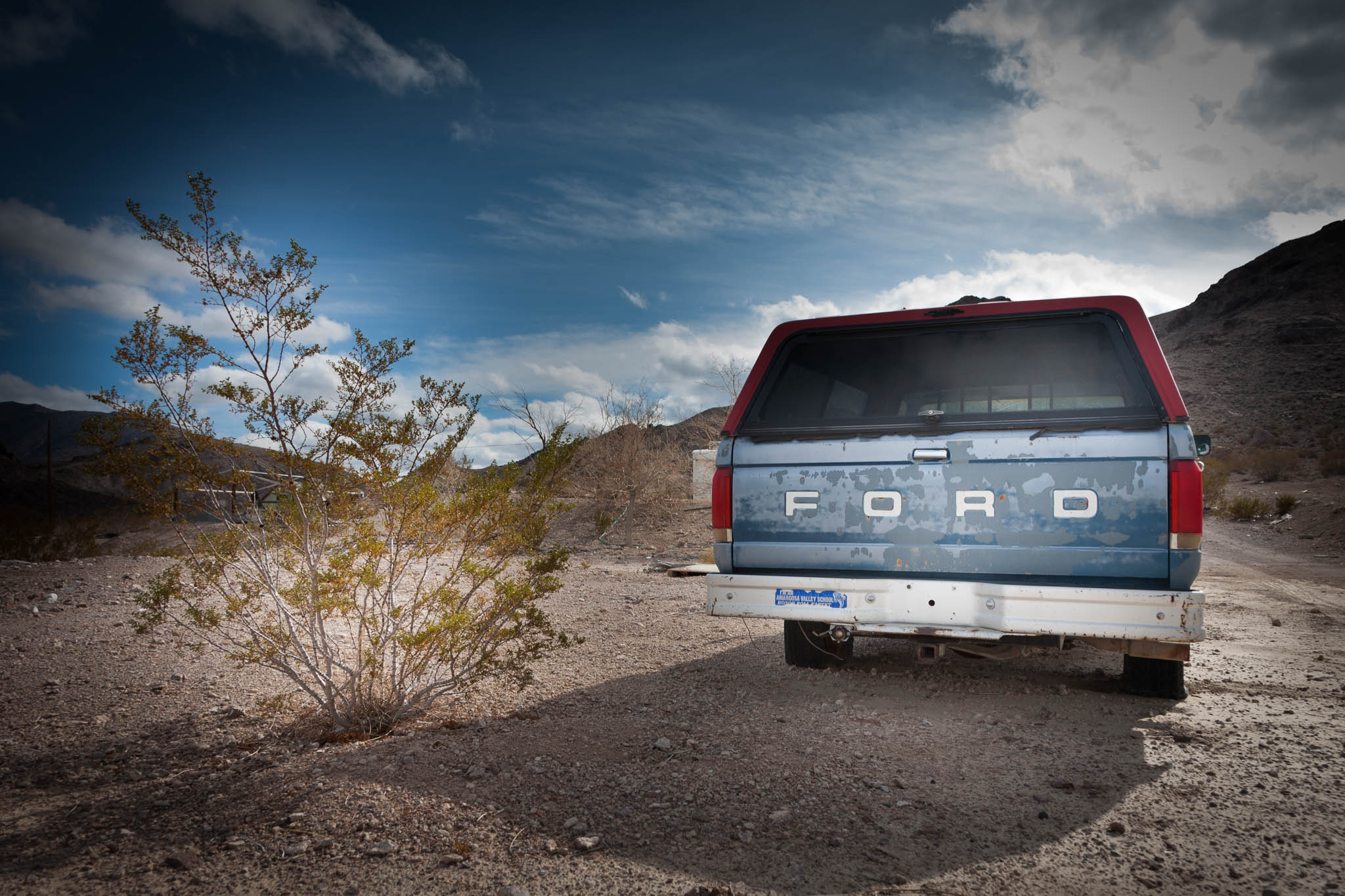 Old blue Ford truck parked on desert dirt road under cloudy sky, with bush and mountains in the background.