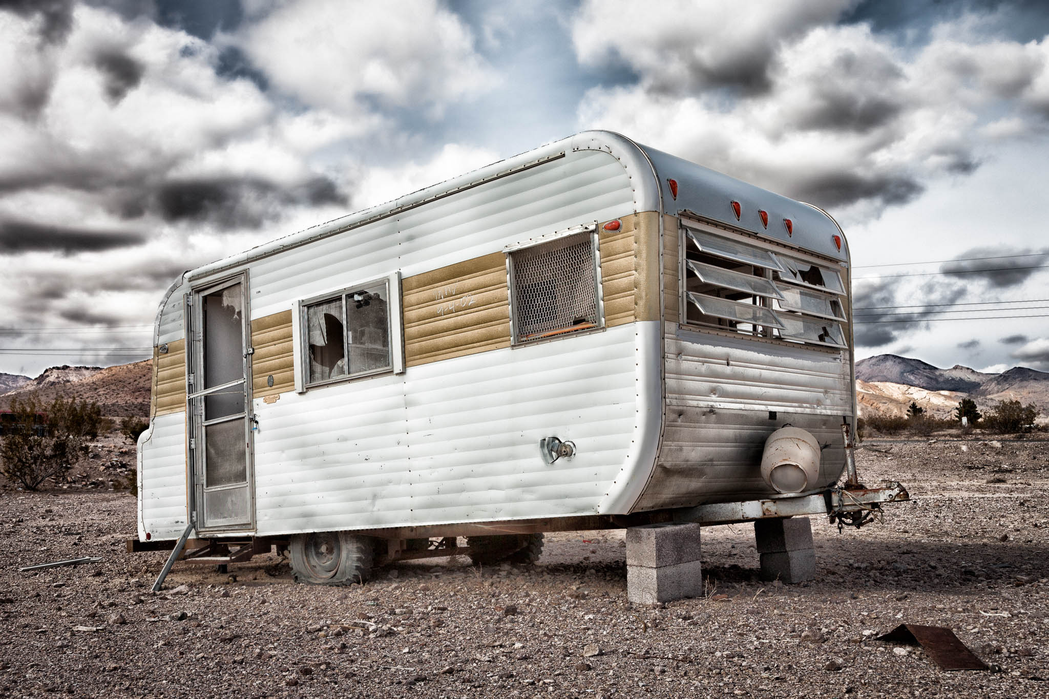 Old camper on blocks in a barren desert landscape under cloudy skies.
