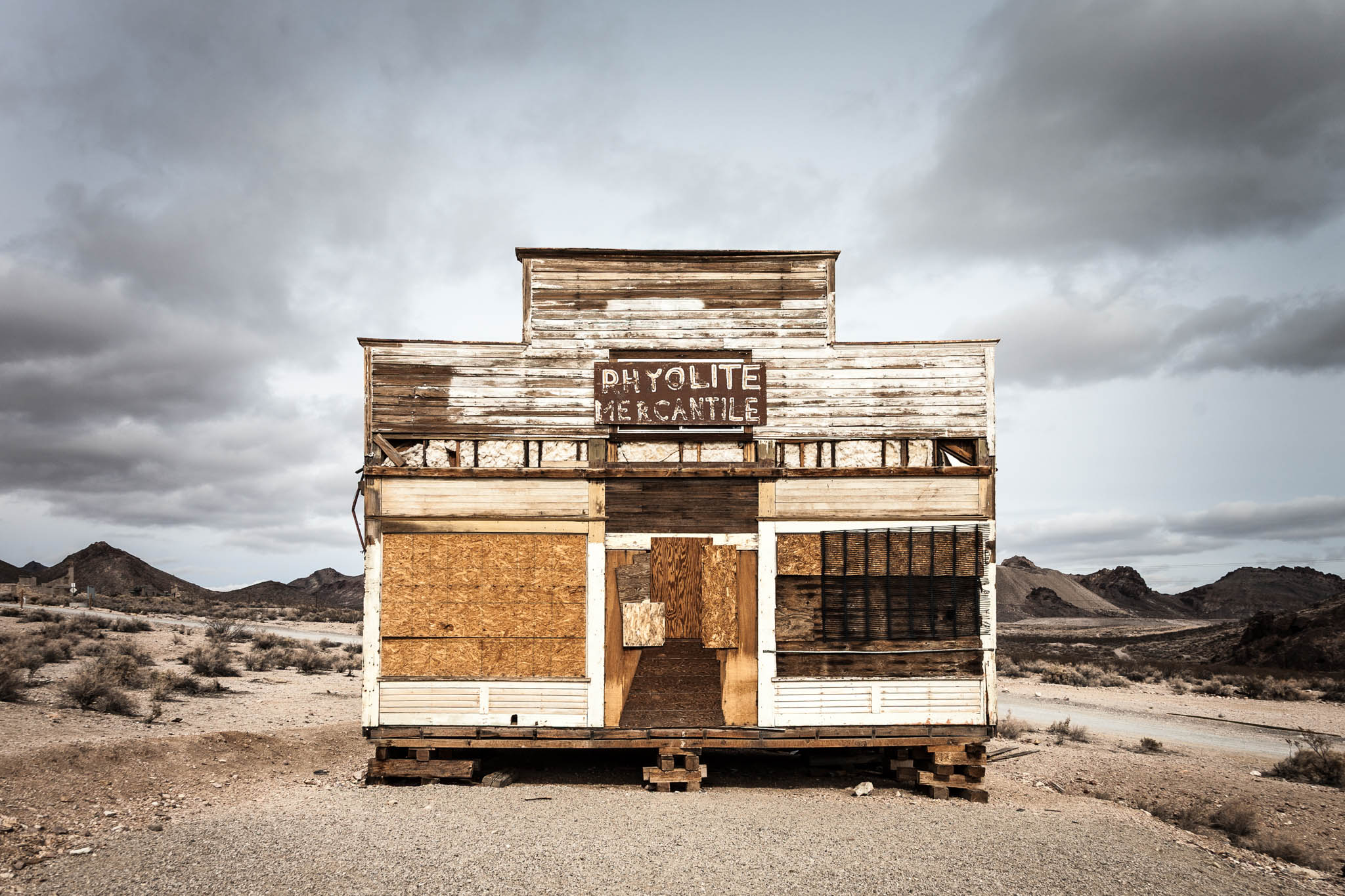 Abandoned Rhyolite Mercantile building in desert ghost town, weathered wood and boarded windows, mountains in background.