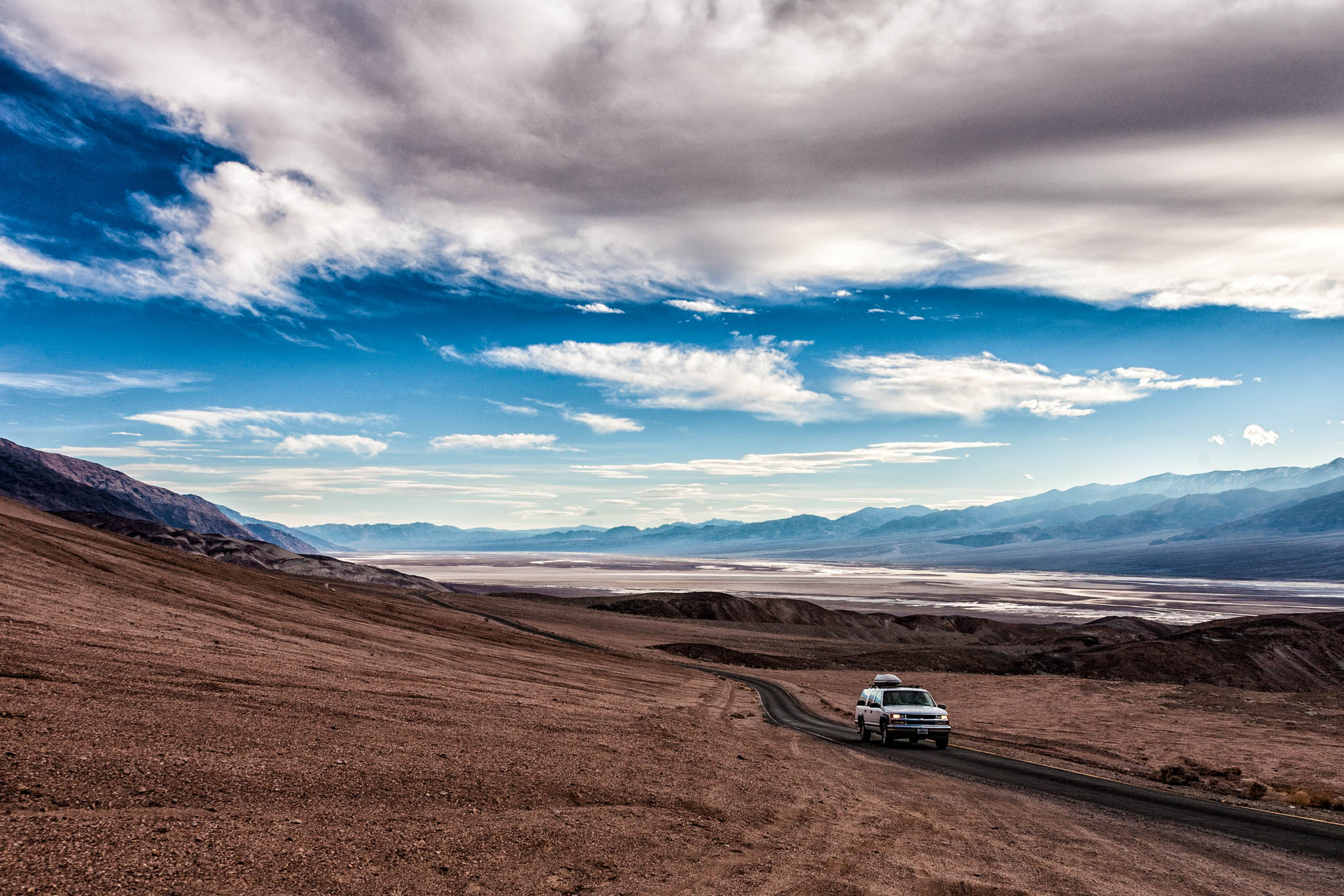 SUV driving on a desert road with expansive mountain views and partly cloudy skies.