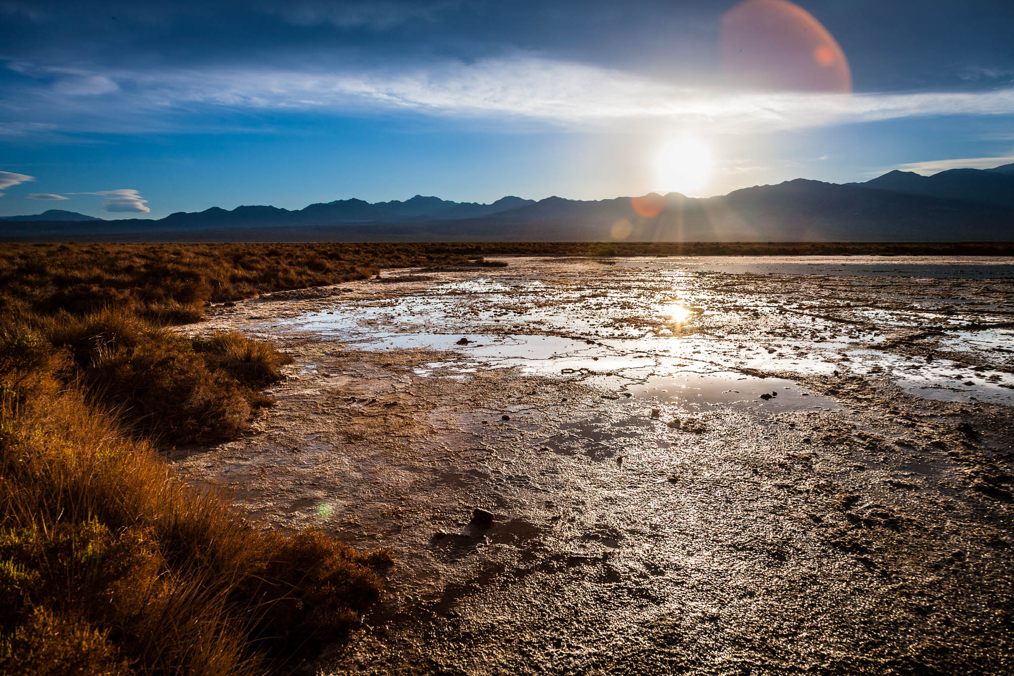 Sunset over a dry, cracked desert landscape with mountains in the distance, under a wide blue sky.