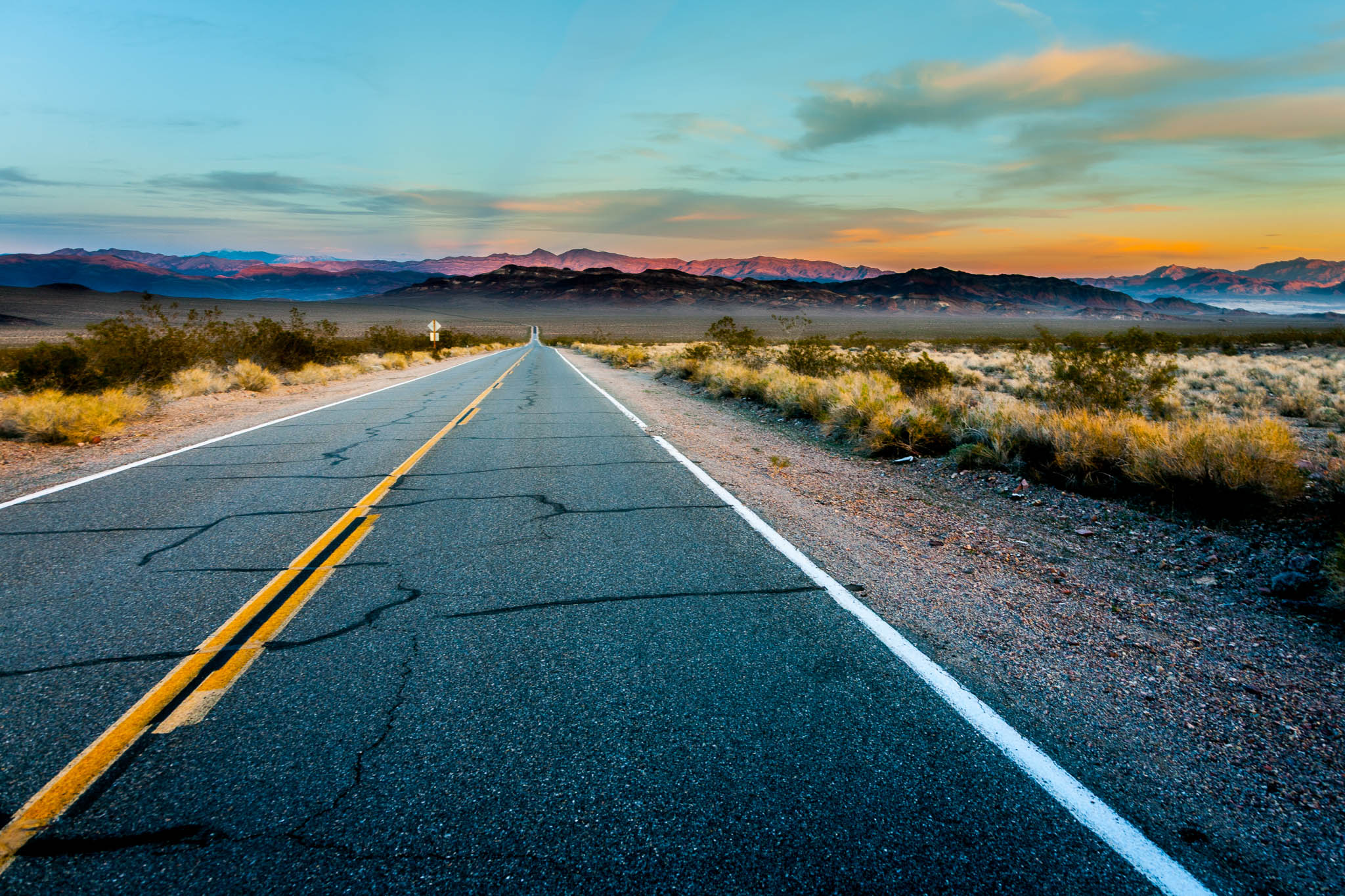Desert highway stretching towards mountains at sunset under a colorful sky, with cracked asphalt and arid landscape.