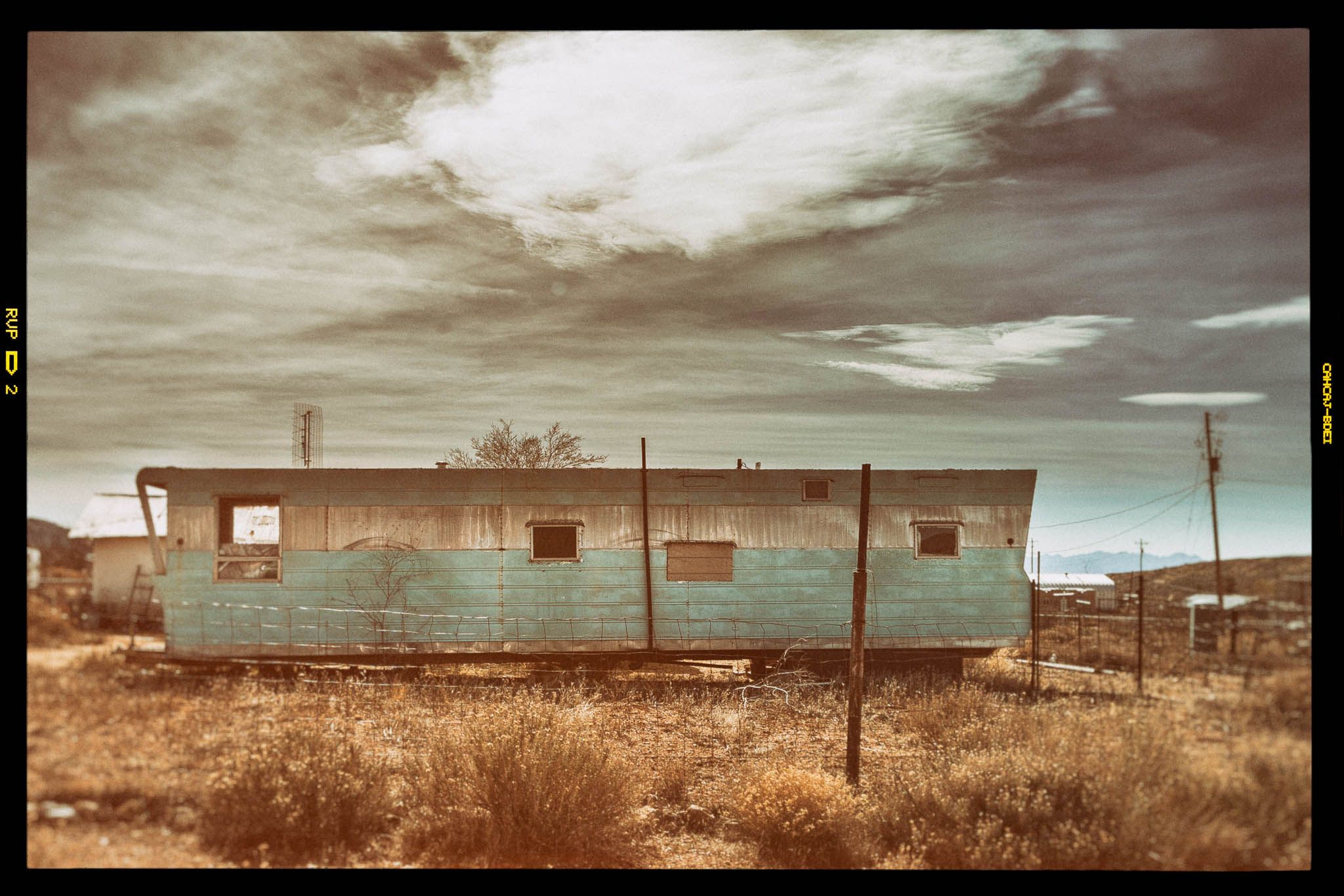 Old blue trailer in a dry field under a cloudy sky, with weathered exterior and surrounding rustic landscape.