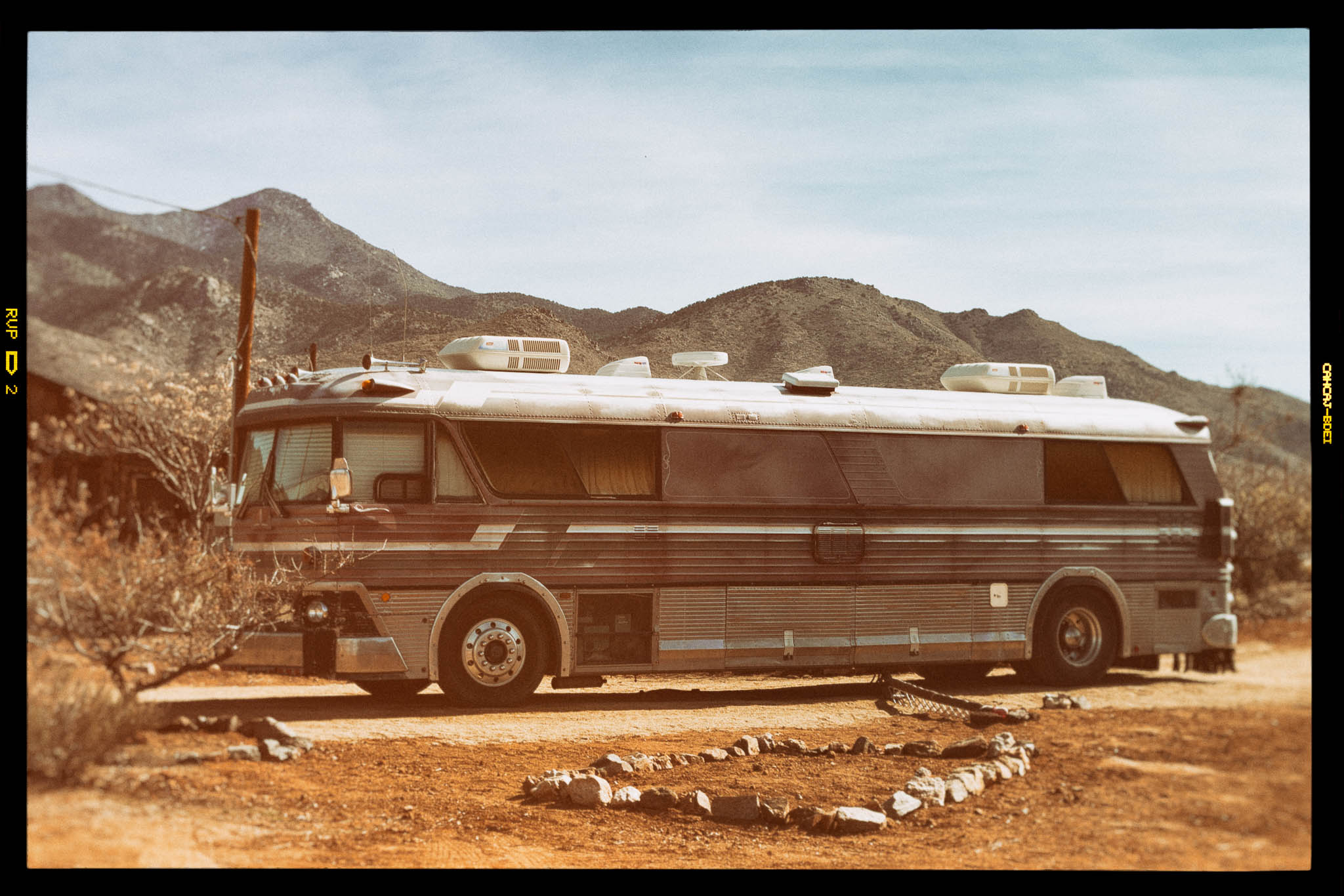 Vintage bus parked on a desert road with mountains in the background, framed by dry shrubs and rocky terrain.