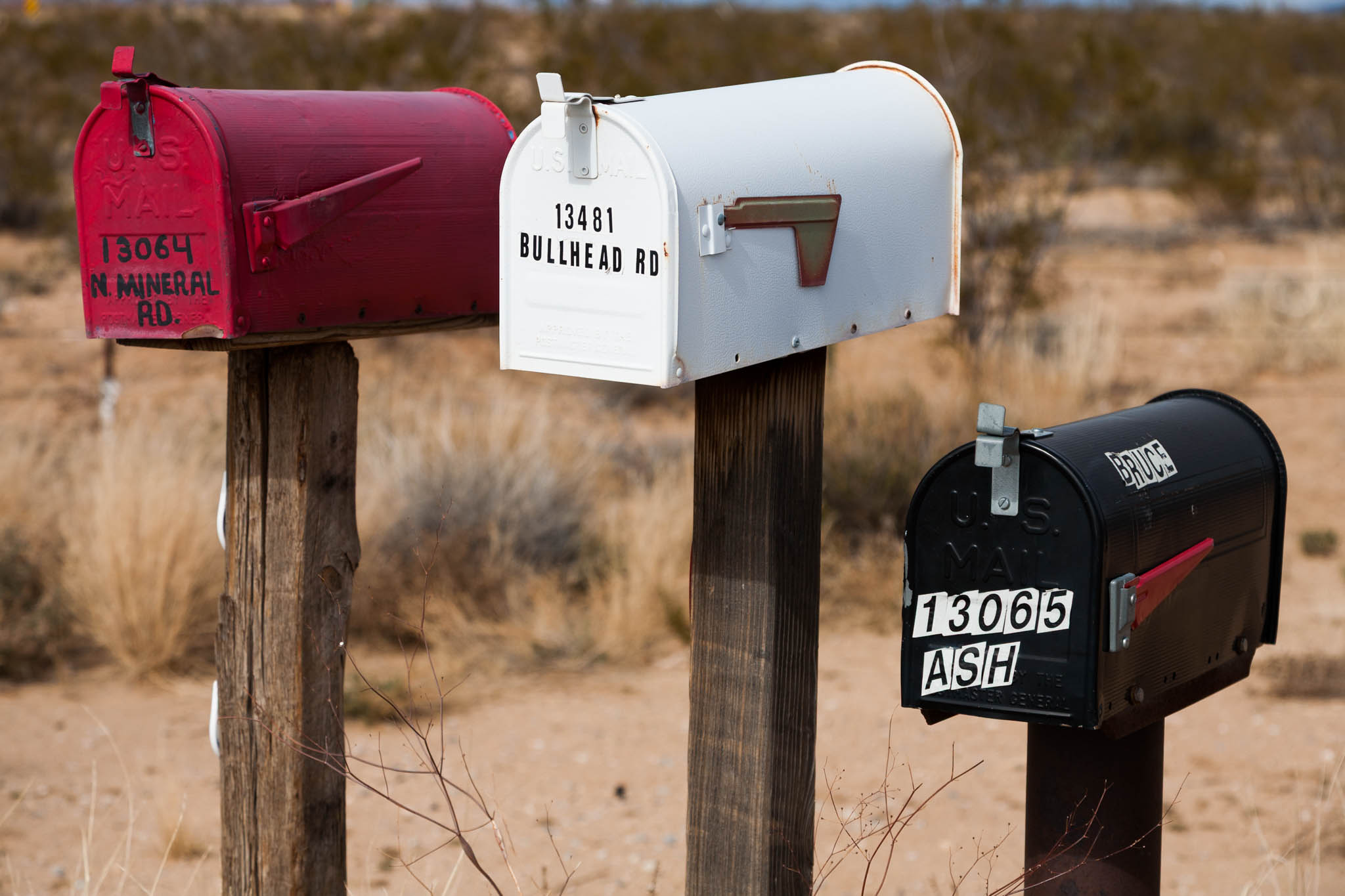 Three mailboxes on wooden posts in a rural desert area, labeled with street addresses.