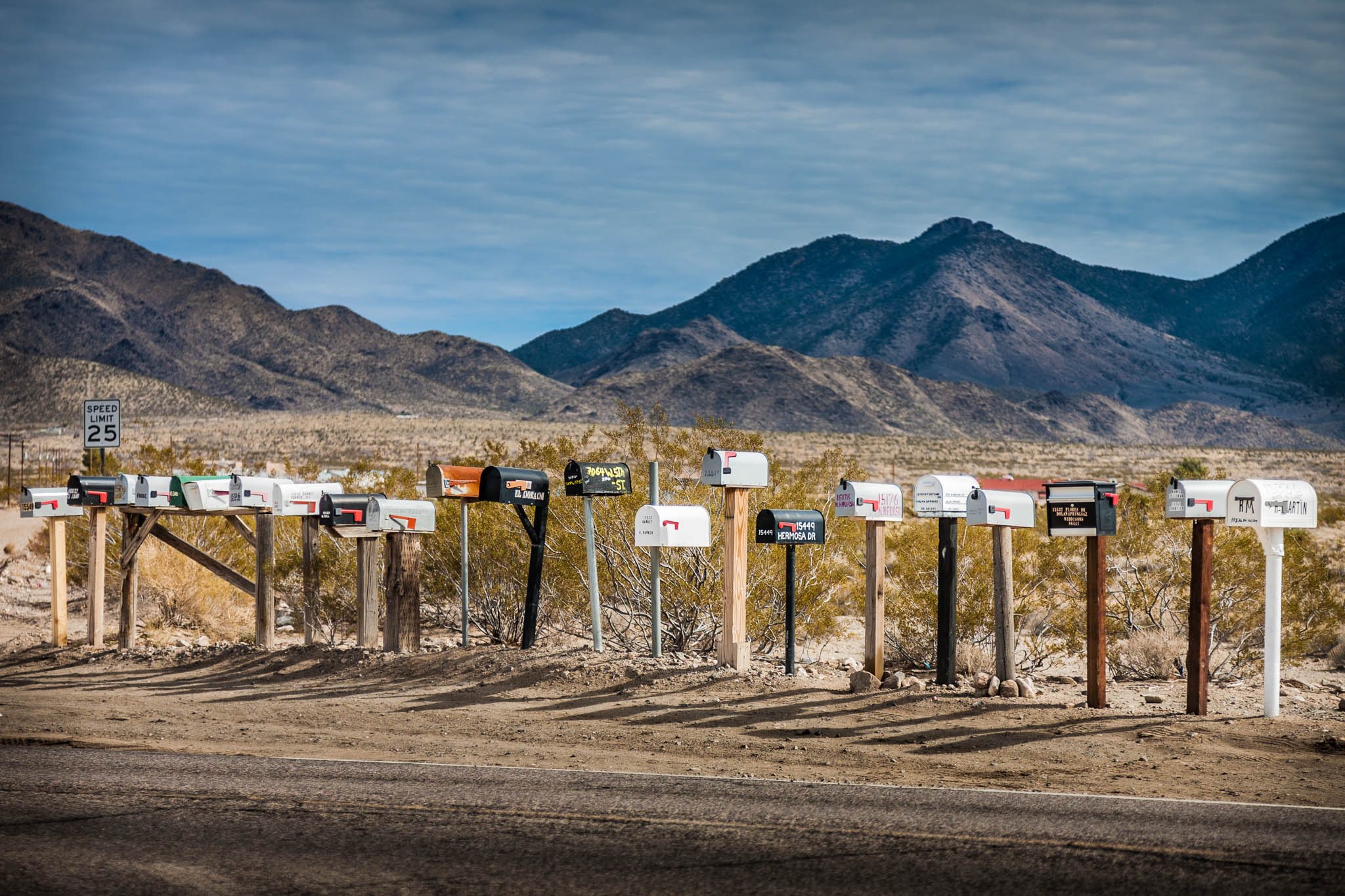 Row of colorful mailboxes on desert roadside with mountains in the background, under a cloudy sky.