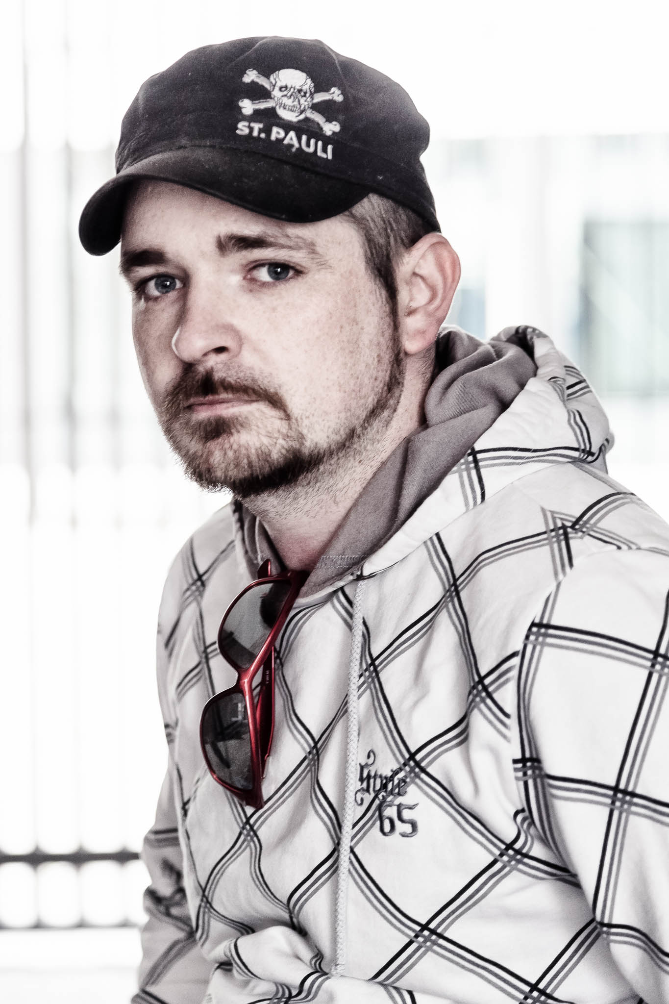 Man wearing a black St. Pauli cap, plaid hoodie, and sunglasses indoors.