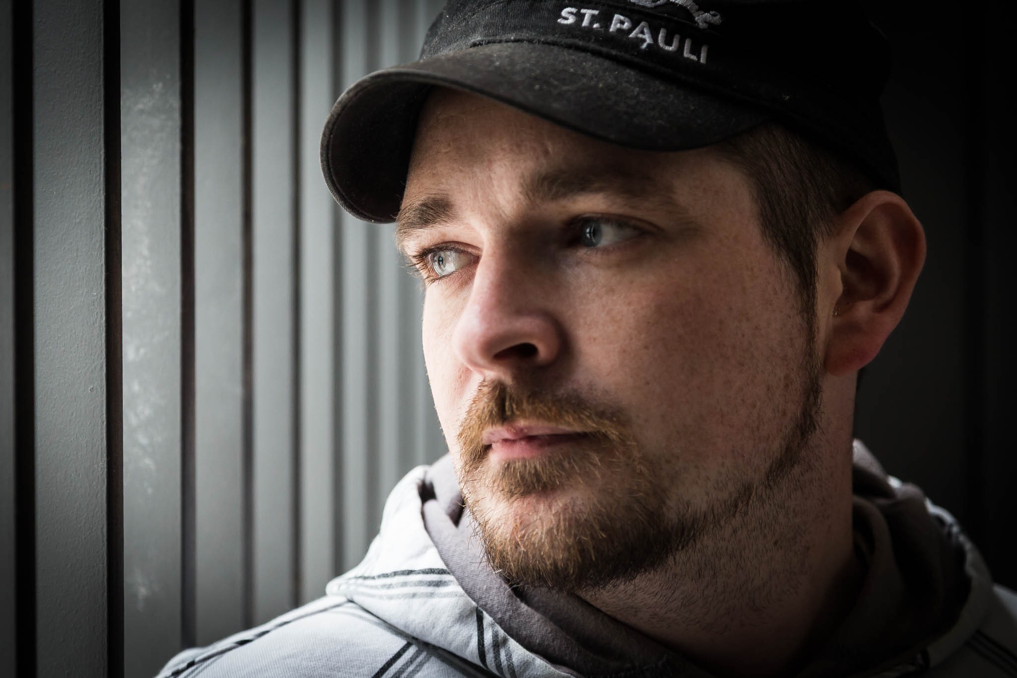 Man in cap looking pensively out a window beside vertical blinds, wearing a hoodie.