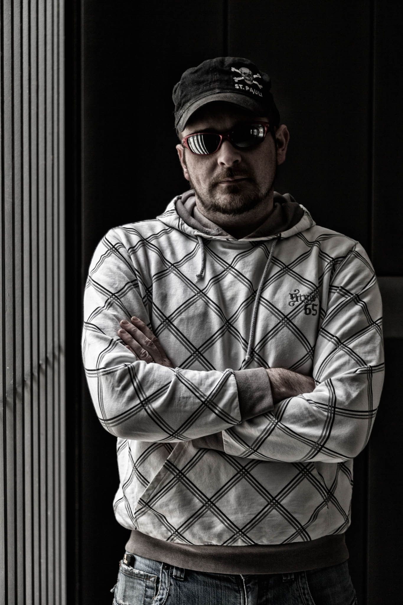 Man wearing sunglasses, St. Pauli cap, and checkered hoodie stands with arms crossed in shadowy, industrial setting.