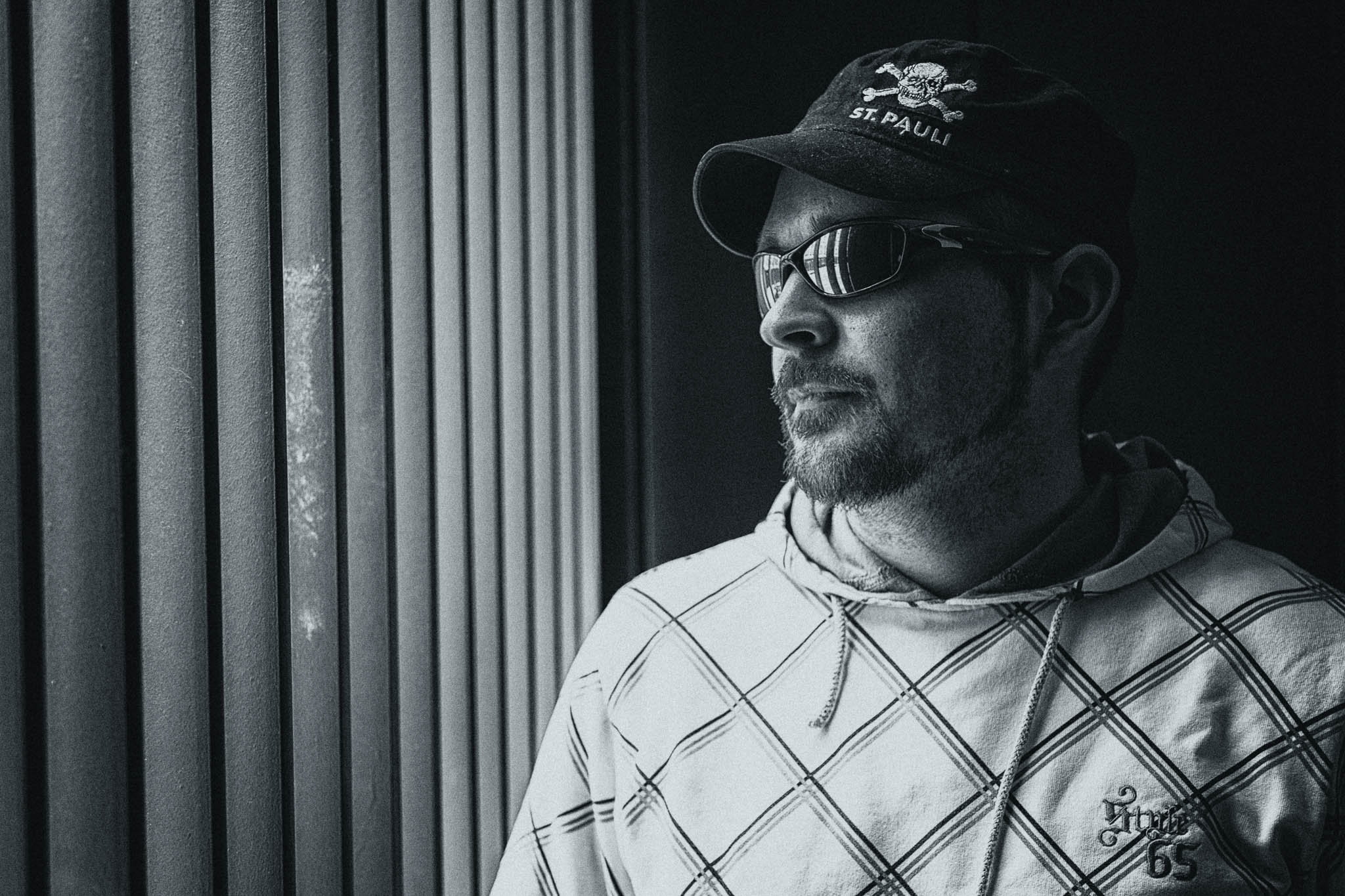 Man in cap and sunglasses gazes outside through window blinds, wearing a patterned hoodie. Black and white photo.