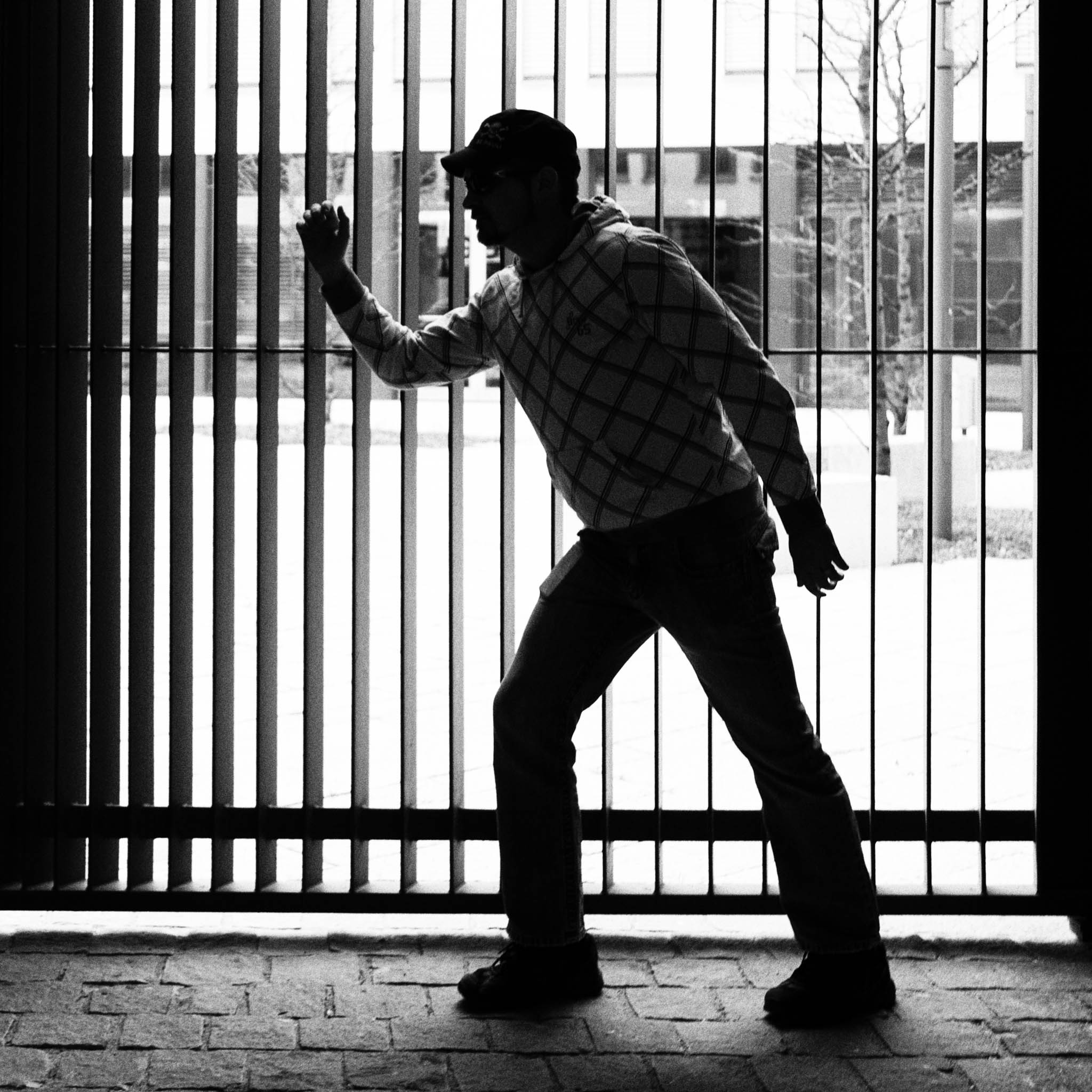 Silhouette of a person in a hoodie and cap walking along vertical metal bars in a black and white scene.