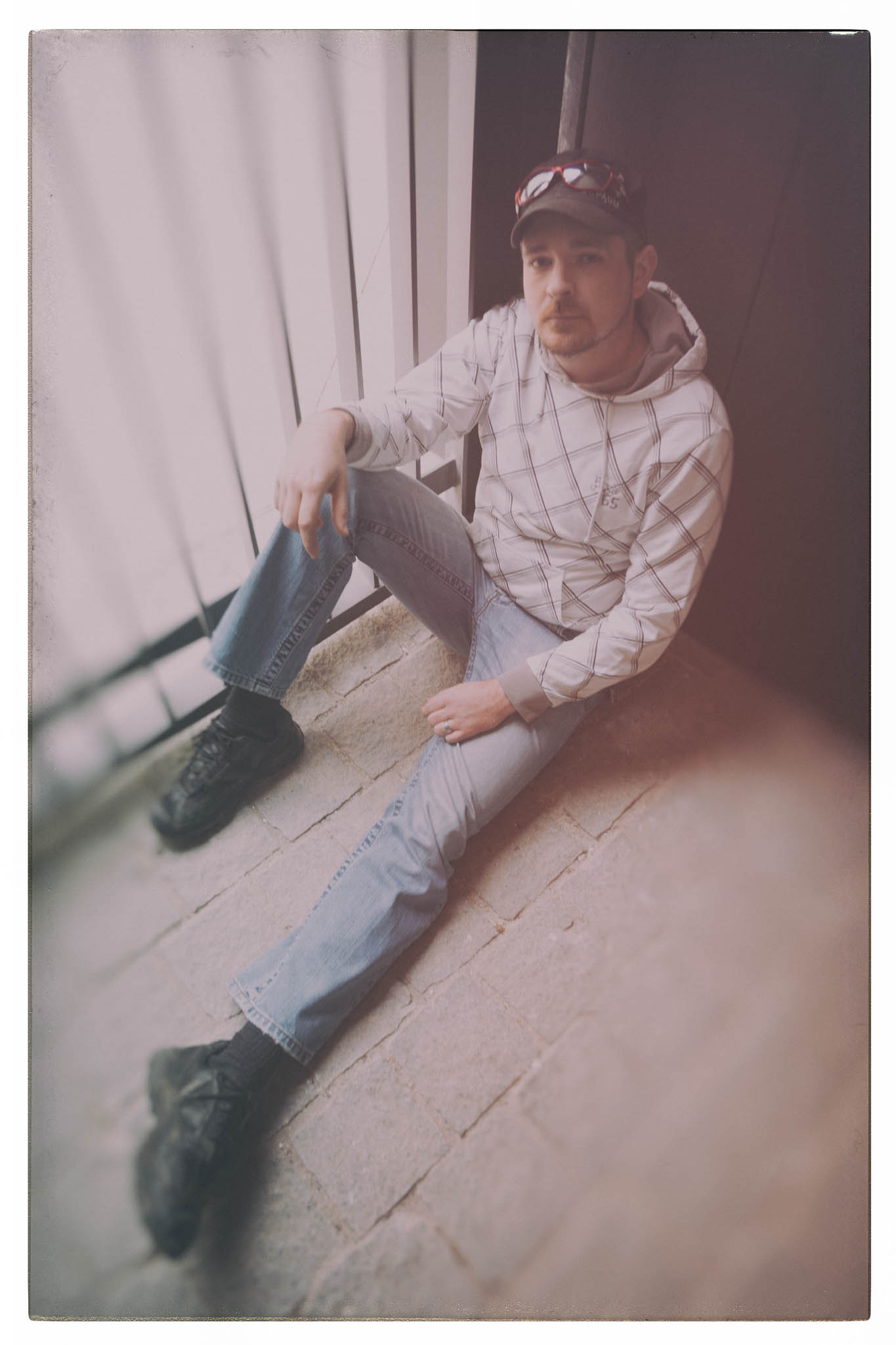 Man in a hoodie and jeans sitting on a brick floor by a railing with a thoughtful expression.