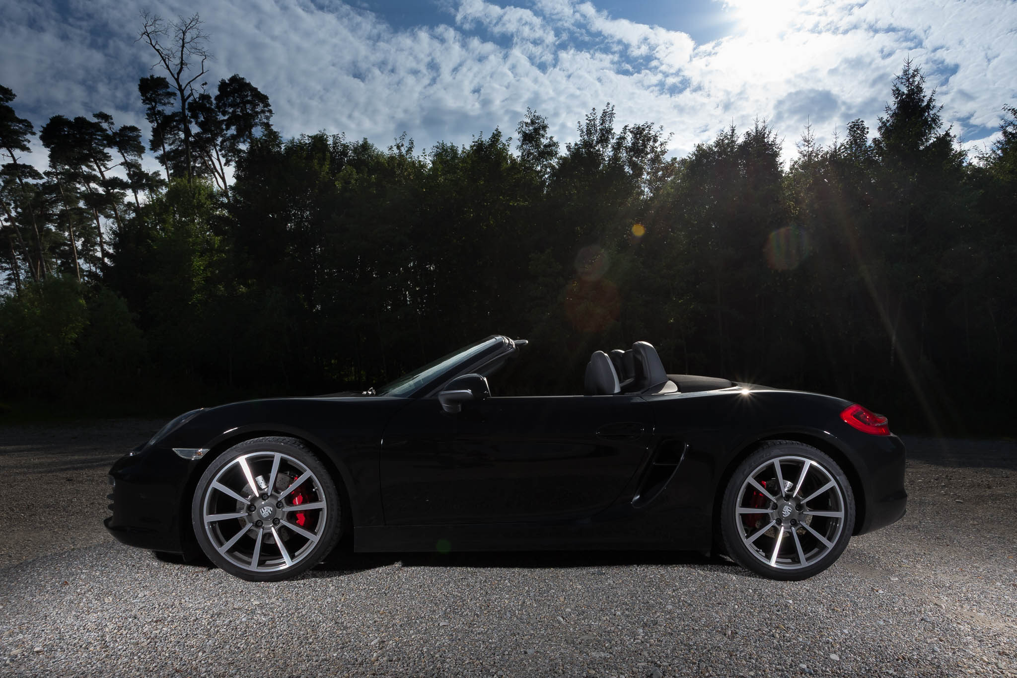 Black convertible sports car parked on gravel, forest background, sunny sky above.