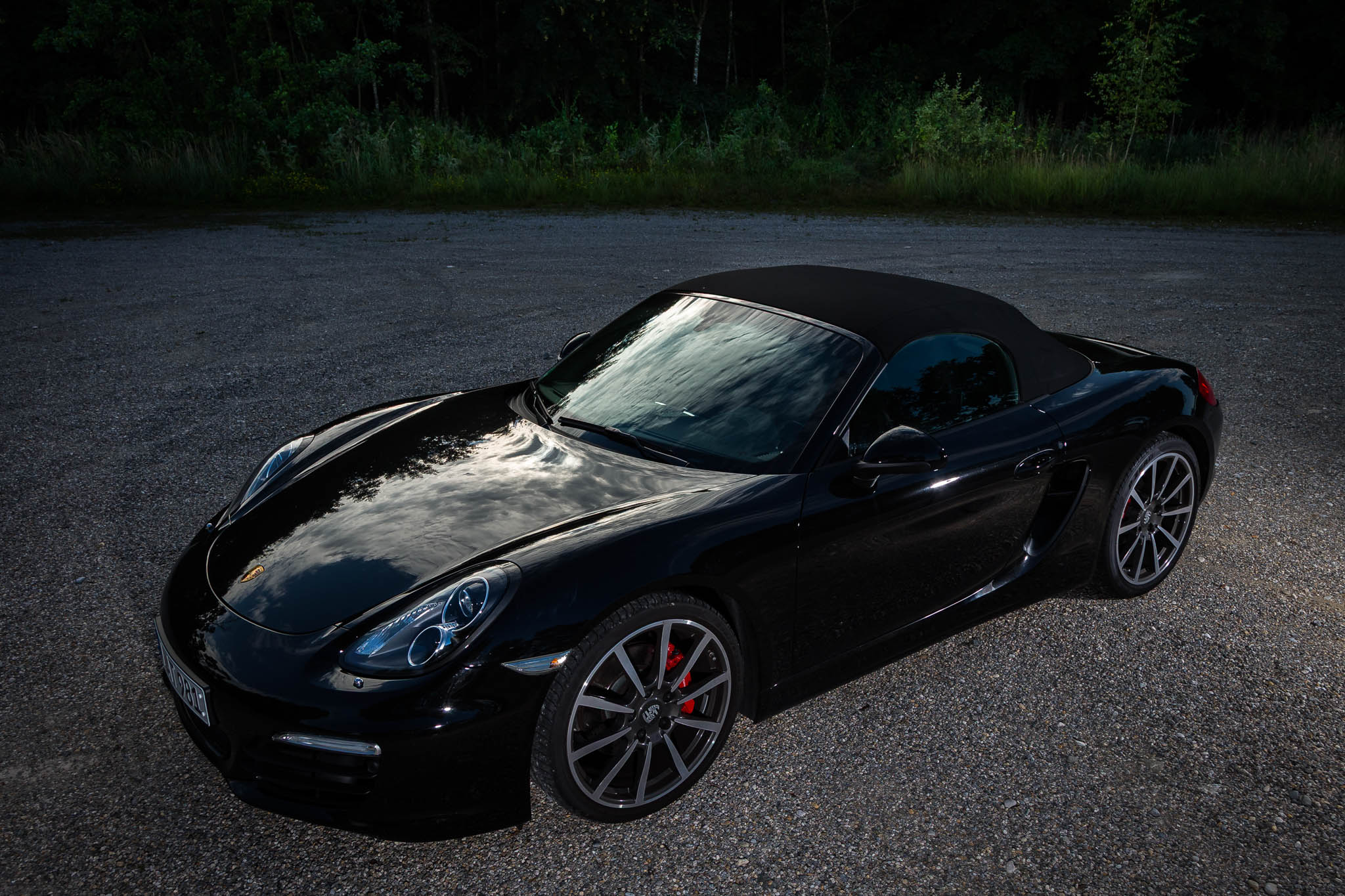Sleek black convertible sports car with reflections of clouds, parked on a gravel lot near lush greenery.