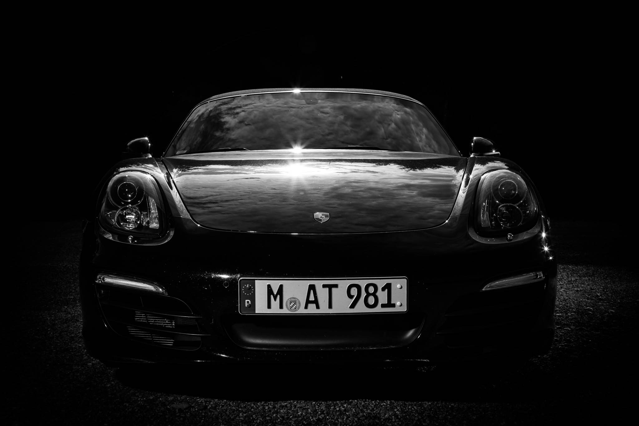 Black sports car with reflective hood, headlights on, and license plate M AT 981 in a dramatic, shadowy setting.