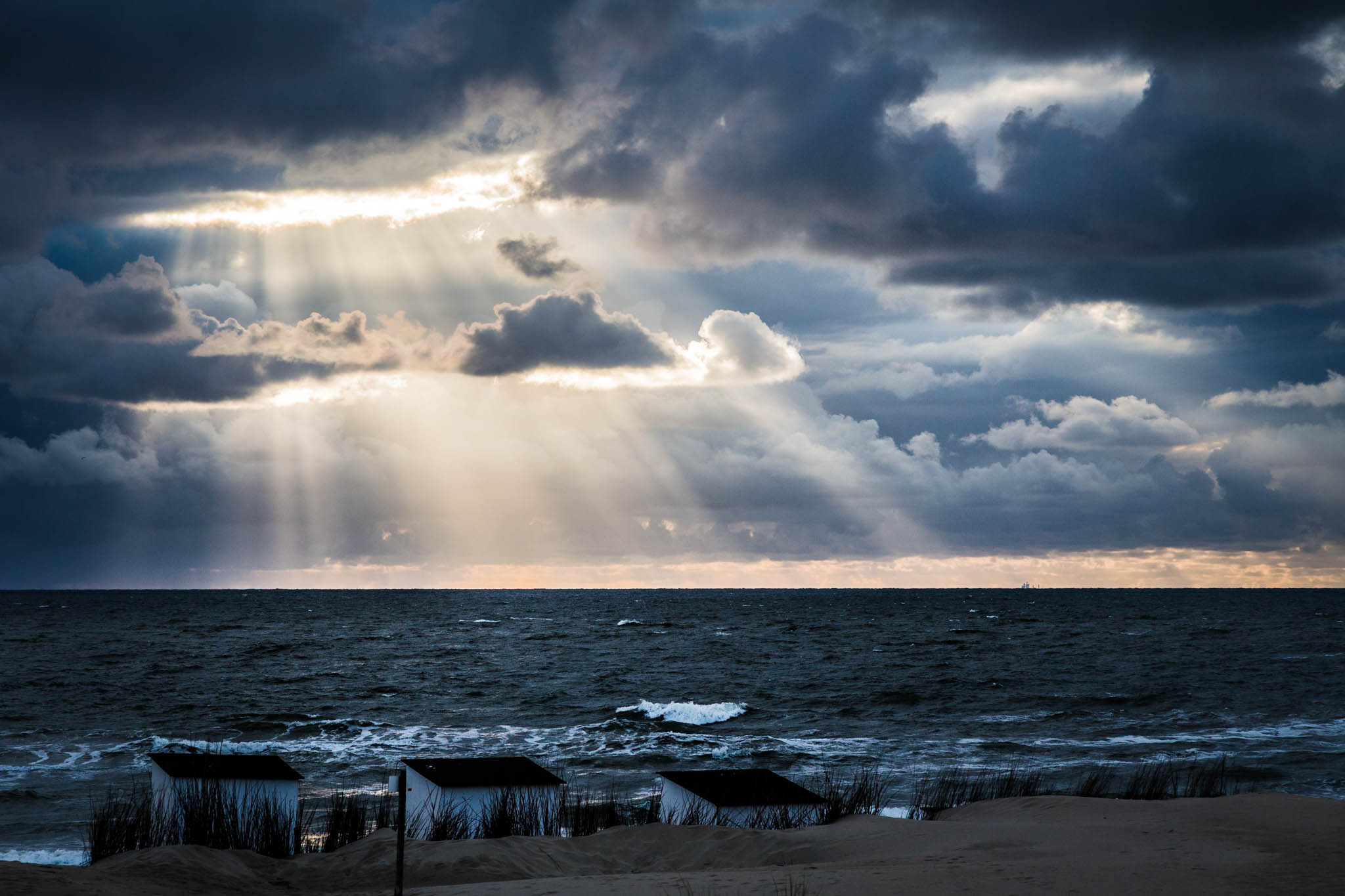 Sun rays break through dramatic clouds over a windy ocean, illuminating beach huts on a sandy shore.