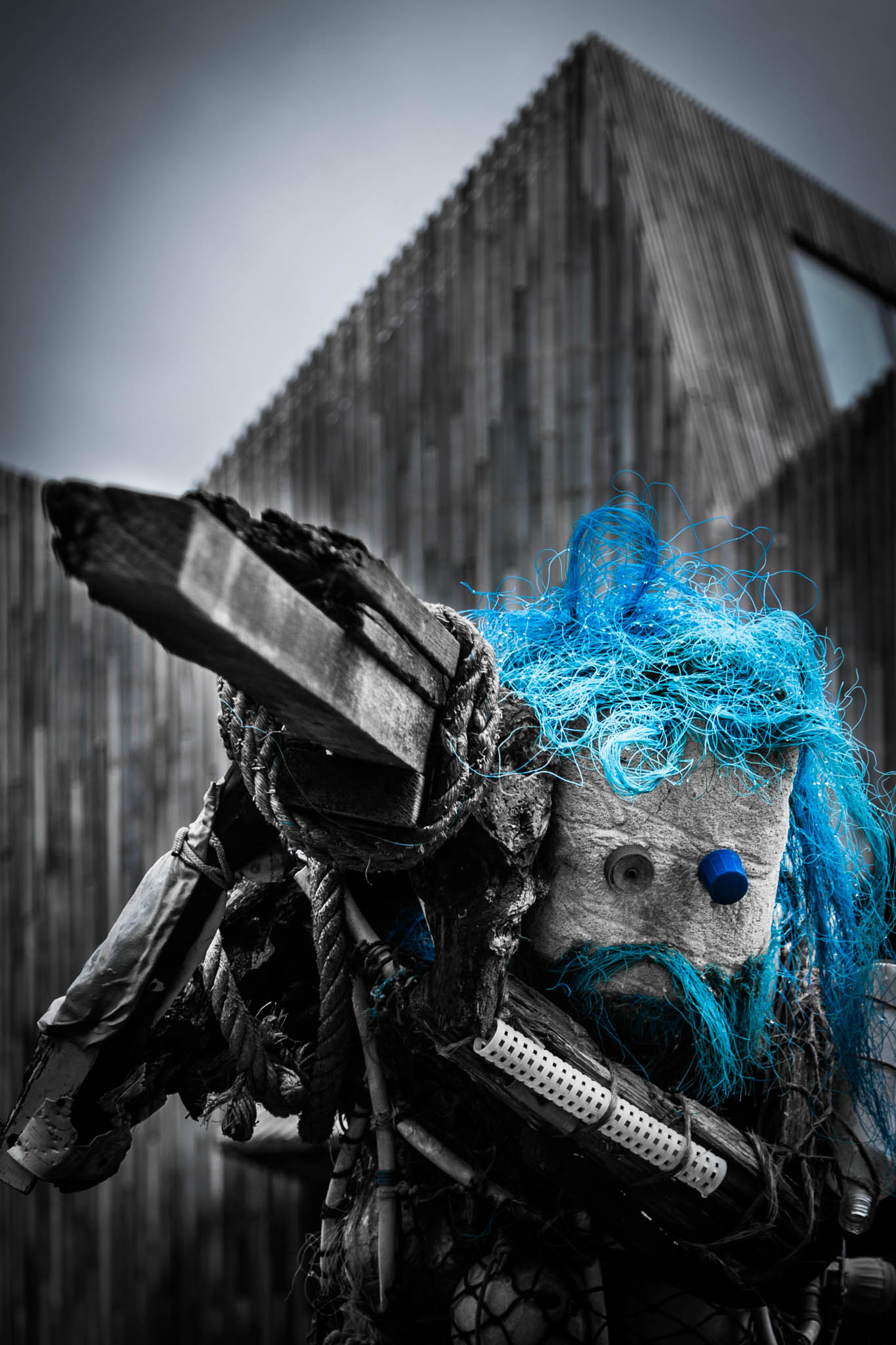 Art sculpture with blue hair made from recycled materials, set against a modern wooden building background.