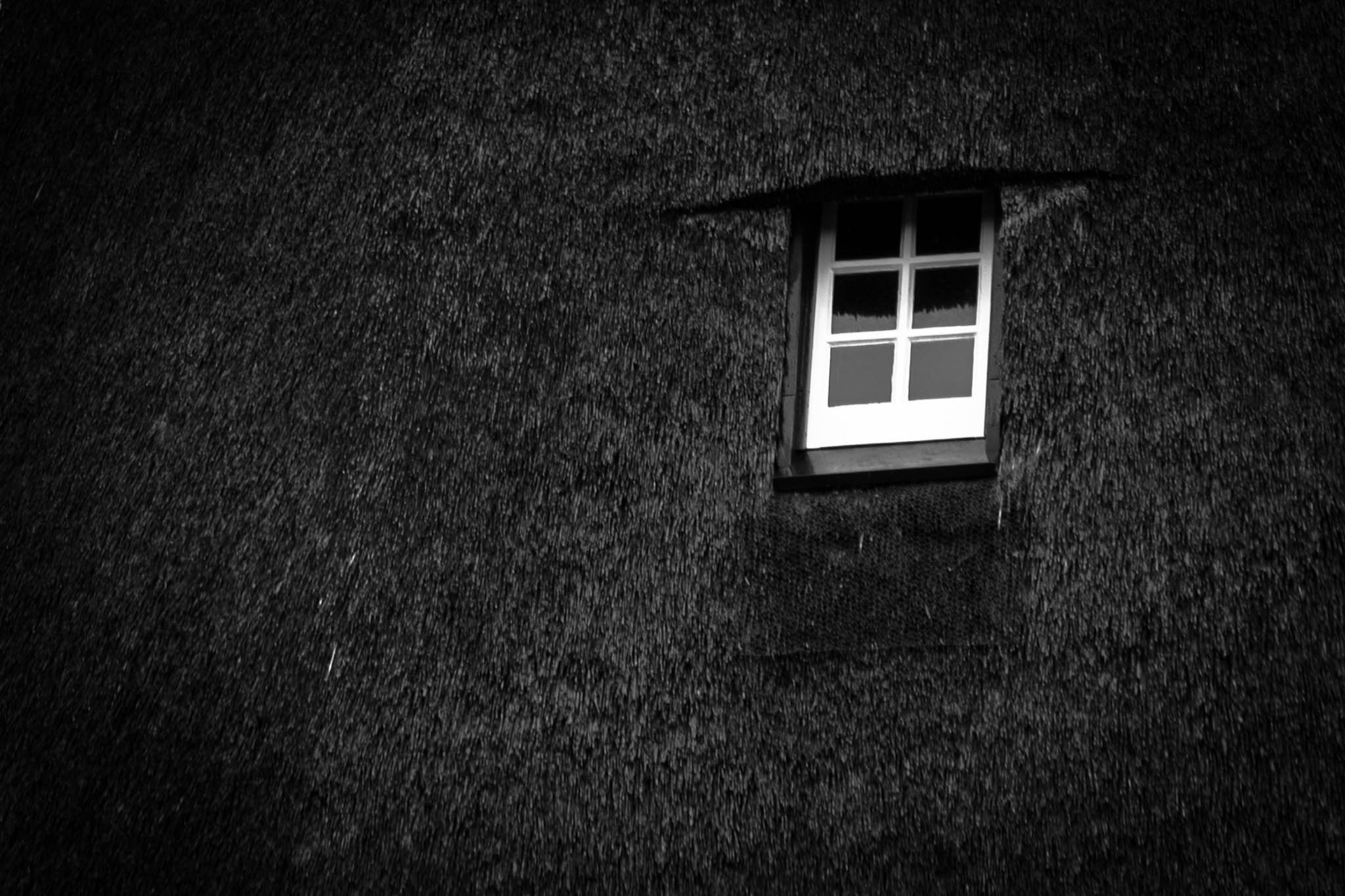 Black and white photo of a small window on a textured dark hut wall.